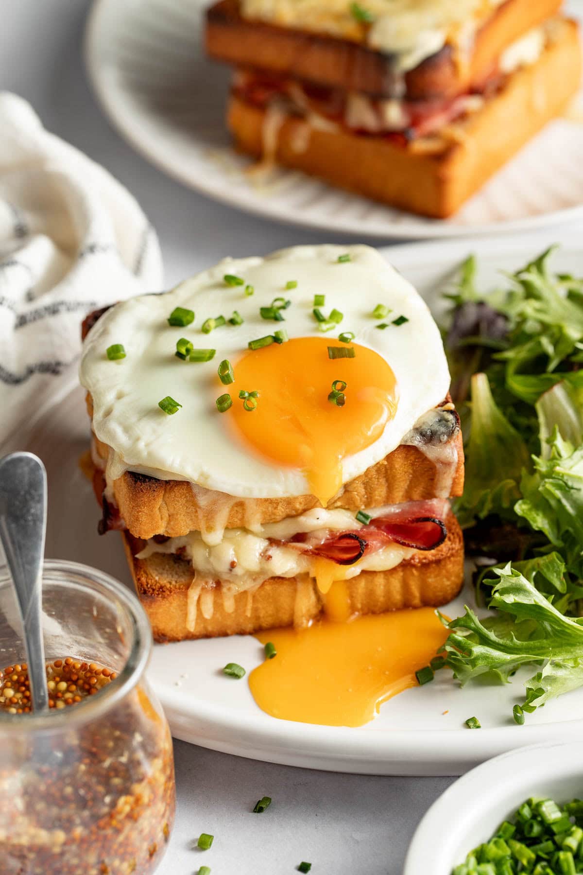 A Croque Madame sandwich topped with a runny fried egg sits on a white plate, with melted cheese and ham between toasted bread. A side of fresh green salad and a jar of grainy mustard are nearby.