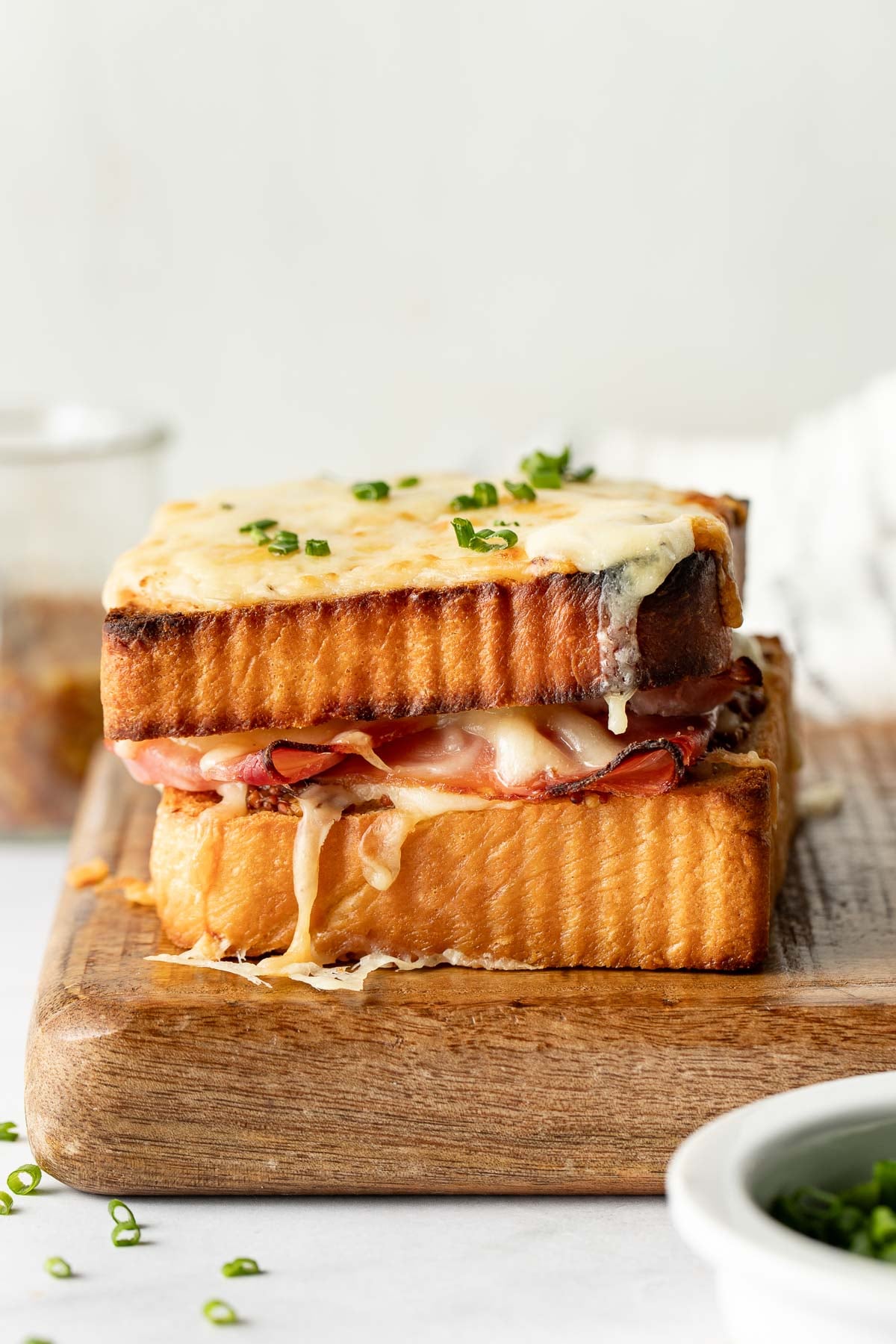 A close-up of a stacked croque monsieur sandwich on a wooden board, with golden toasted bread, melted cheese, and slices of ham, garnished with chopped chives.