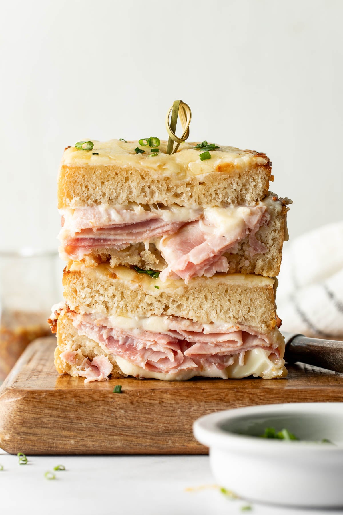 A close-up of a stacked ham and cheese sandwich, with melted cheese and creamy sauce, served on a wooden board and garnished with chopped chives.