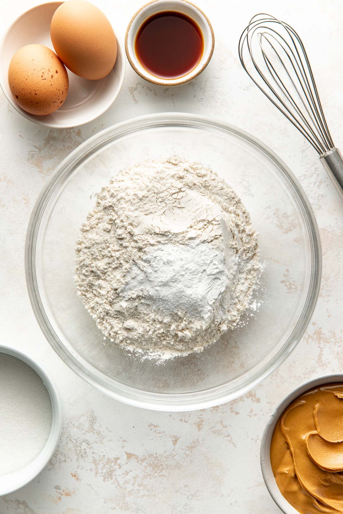 A glass bowl of flour with baking powder sits on a light surface, surrounded by a whisk, two eggs in a dish, a bowl of vanilla extract, a bowl of sugar, and a bowl of peanut butter.