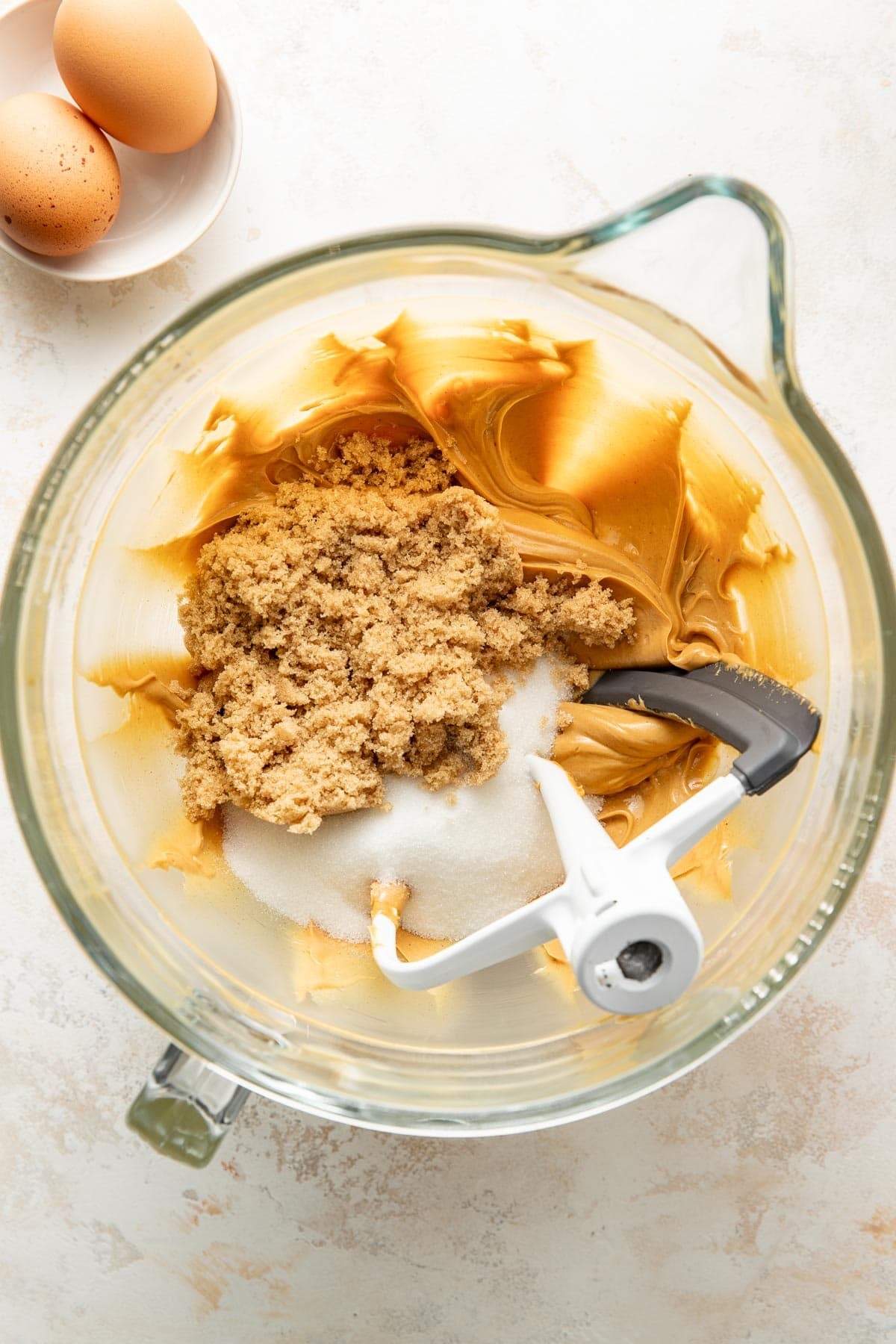 A mixing bowl with brown sugar, white sugar, and creamy peanut butter being mixed, with two eggs in a small bowl nearby on a light countertop.