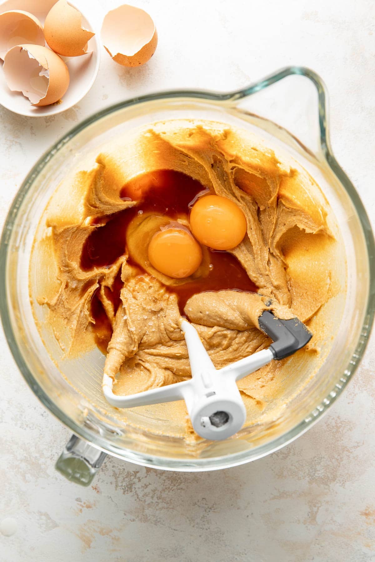 A mixing bowl with creamed ingredients, two eggs, and vanilla extract, with a stand mixer paddle attachment. Broken eggshells are in a small bowl nearby on a light countertop.