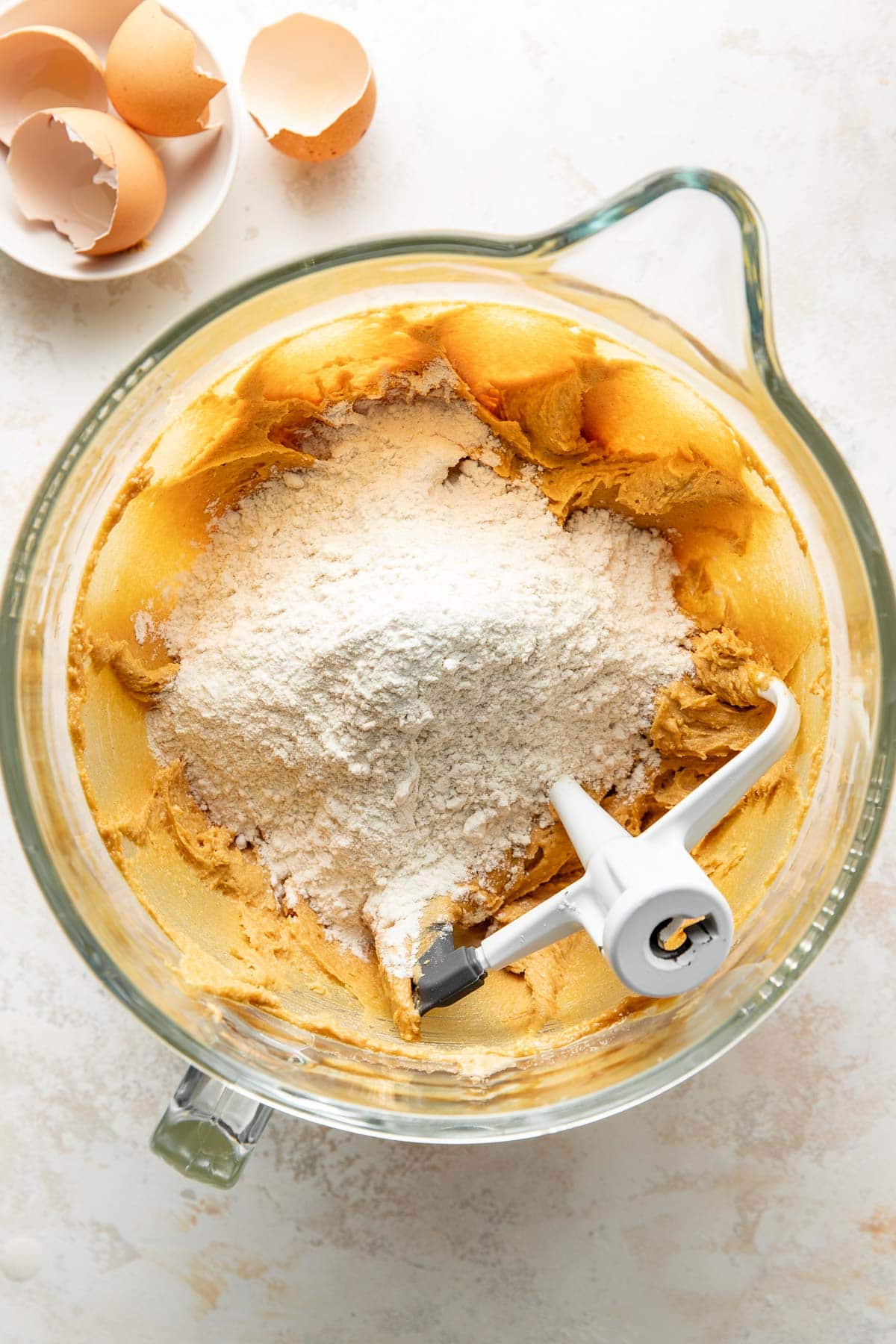 A glass mixing bowl containing creamed butter and sugar with flour on top, partially mixed with a stand mixer attachment. Broken eggshells are in a small bowl nearby on a light countertop.