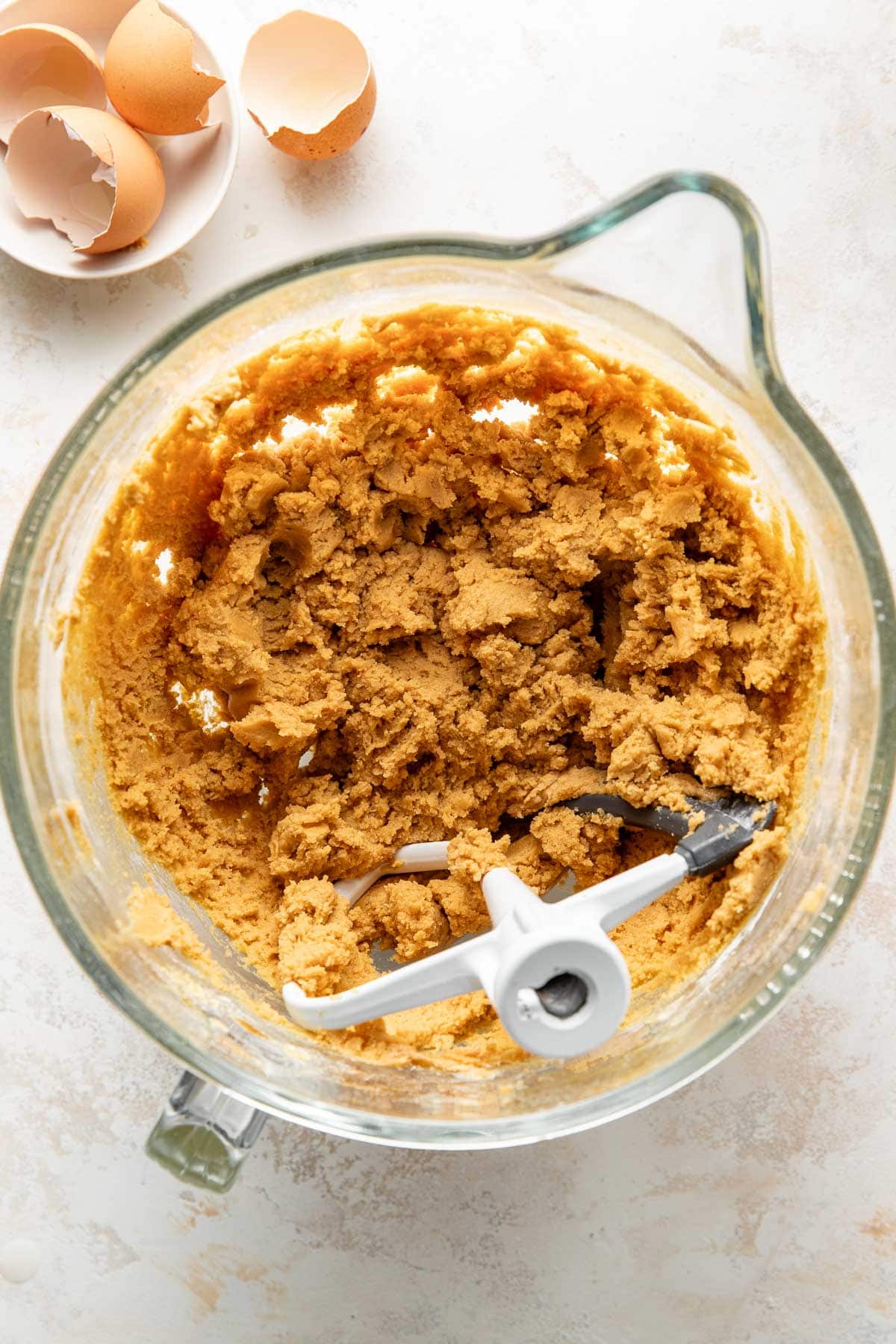 A glass mixing bowl filled with cookie dough next to cracked egg shells on a light-colored surface. A paddle attachment is inside the bowl, indicating the dough has been freshly mixed.