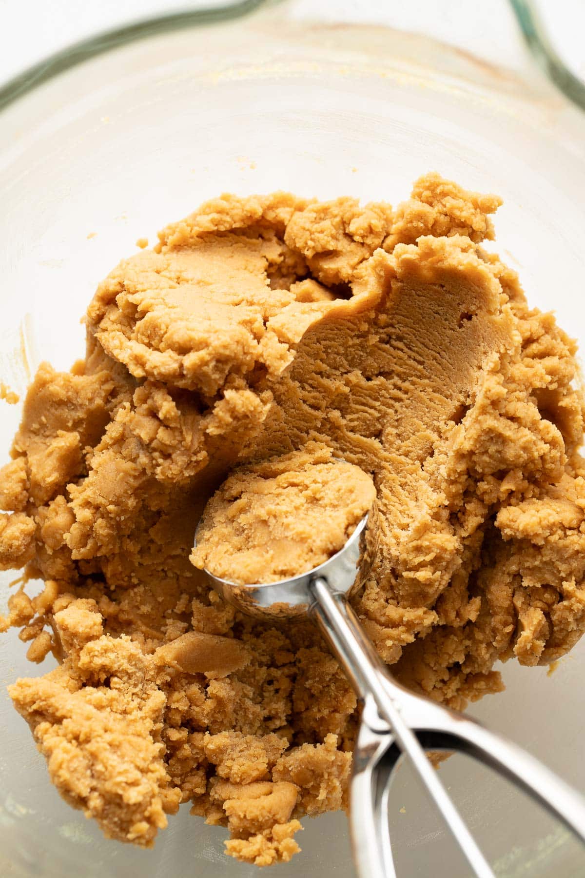 A metal cookie scoop is pressing into a ball of peanut butter cookie dough in a glass mixing bowl. The dough appears creamy and slightly crumbly in texture.