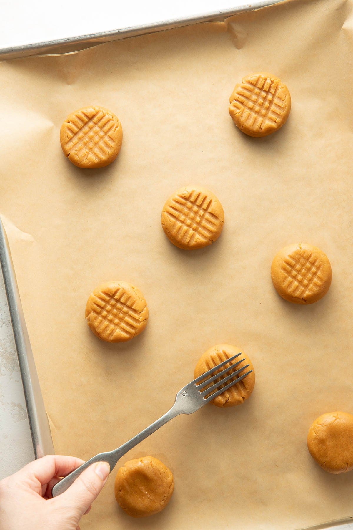 A hand uses a fork to press a crisscross pattern onto round peanut butter cookie dough balls on a baking sheet lined with parchment paper.