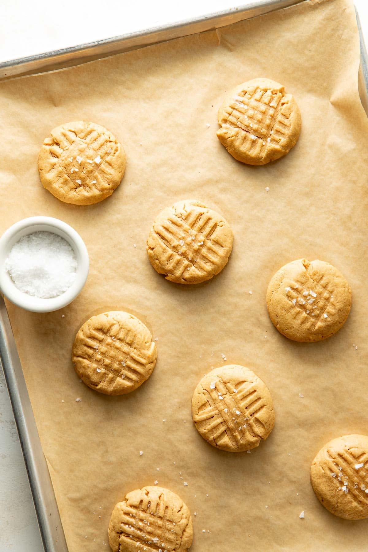 A baking sheet lined with parchment paper holds eight peanut butter cookies with crisscross fork marks, some sprinkled with salt. A small bowl of coarse salt sits in the top left corner.