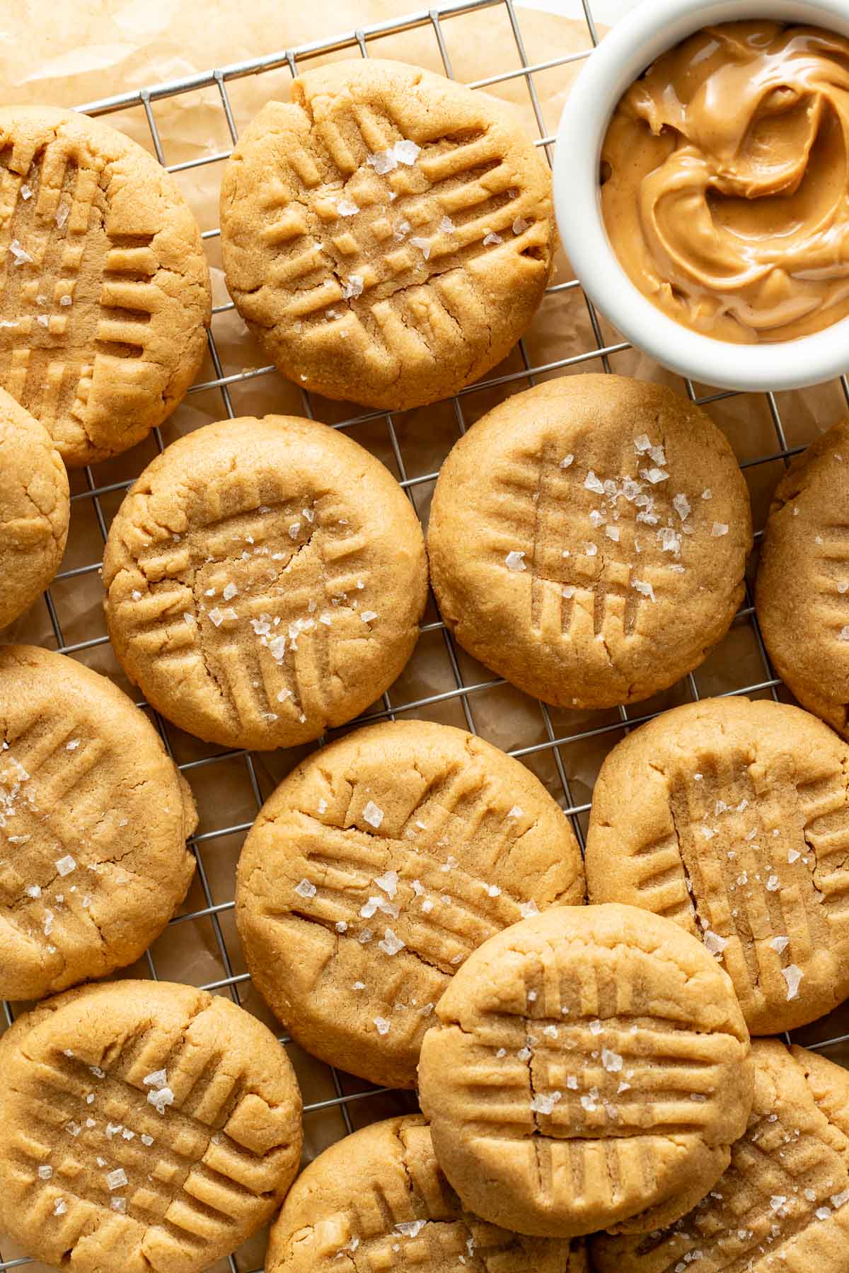 Peanut butter cookies with fork marks and a sprinkle of sea salt are cooling on a wire rack next to a small bowl of creamy peanut butter.