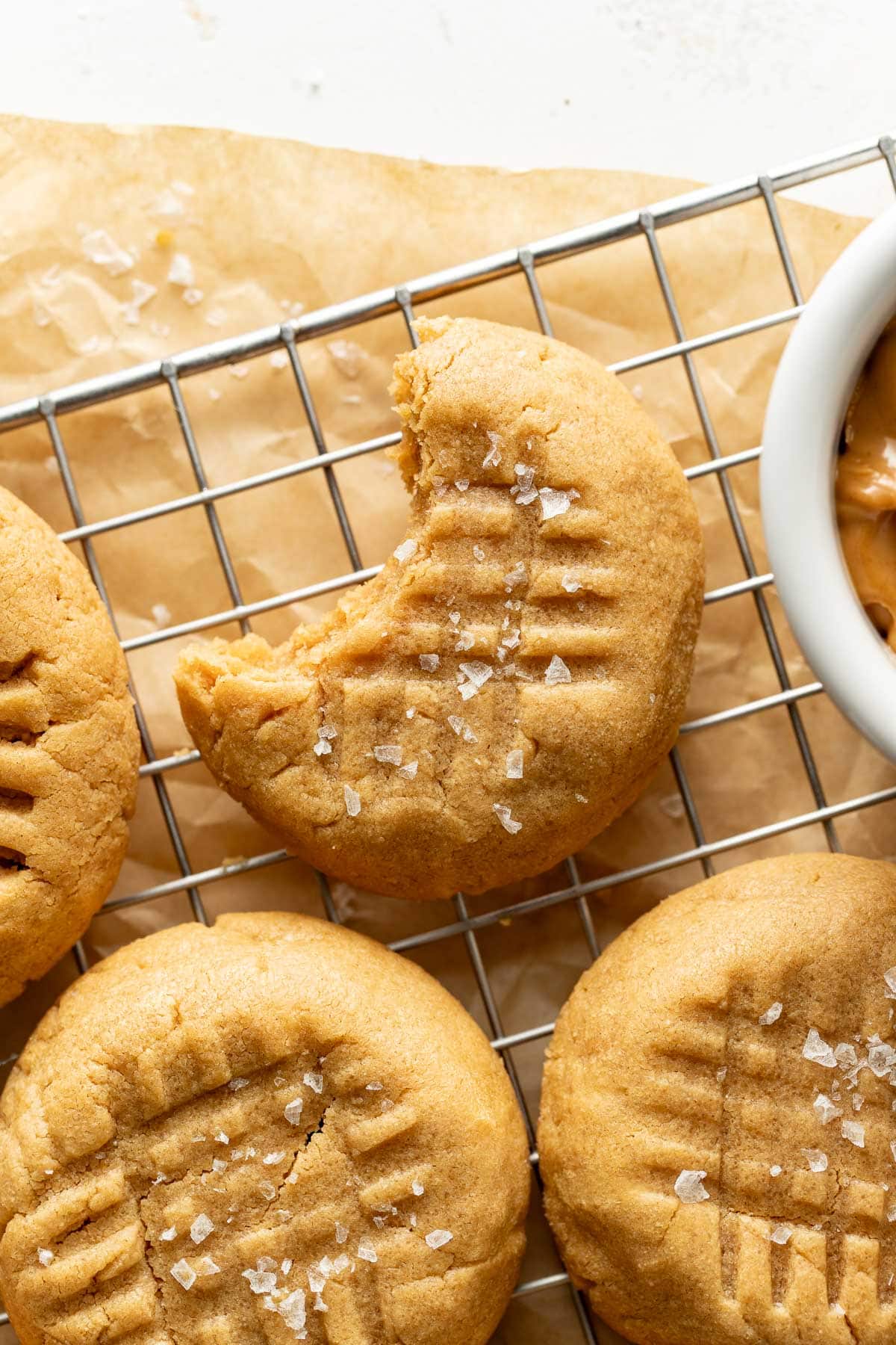Three peanut butter cookies topped with flaky sea salt sit on a wire cooling rack. One cookie has a bite taken out of it. A small dish with peanut butter is partially visible on the right.