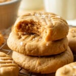 Three peanut butter cookies stacked on a cooling rack, with the top cookie showing a bite taken out. The cookies are sprinkled with coarse salt and there are more cookies and a glass of milk in the background.