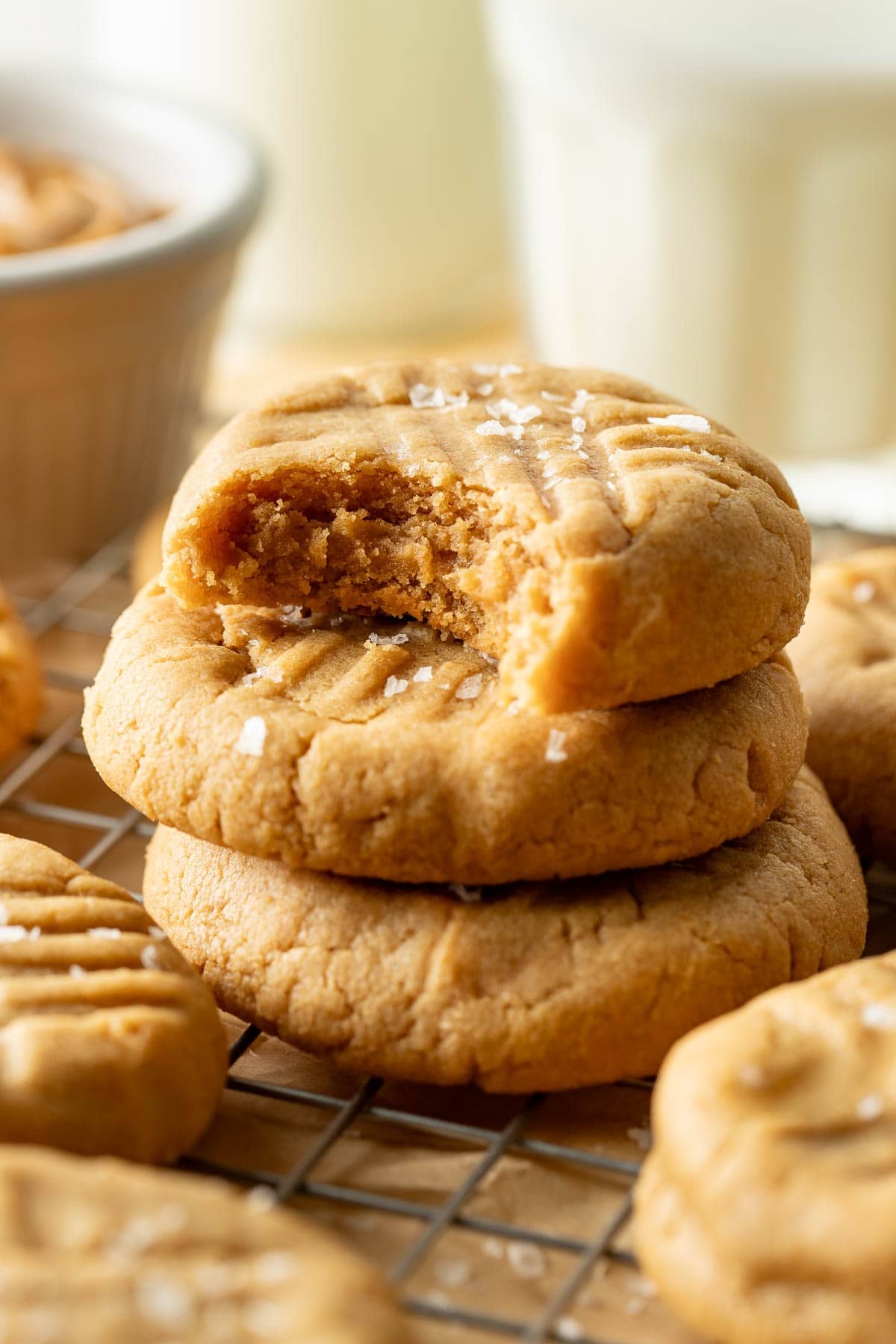Three peanut butter cookies stacked on a cooling rack, with the top cookie showing a bite taken out. The cookies are sprinkled with coarse salt and there are more cookies and a glass of milk in the background.