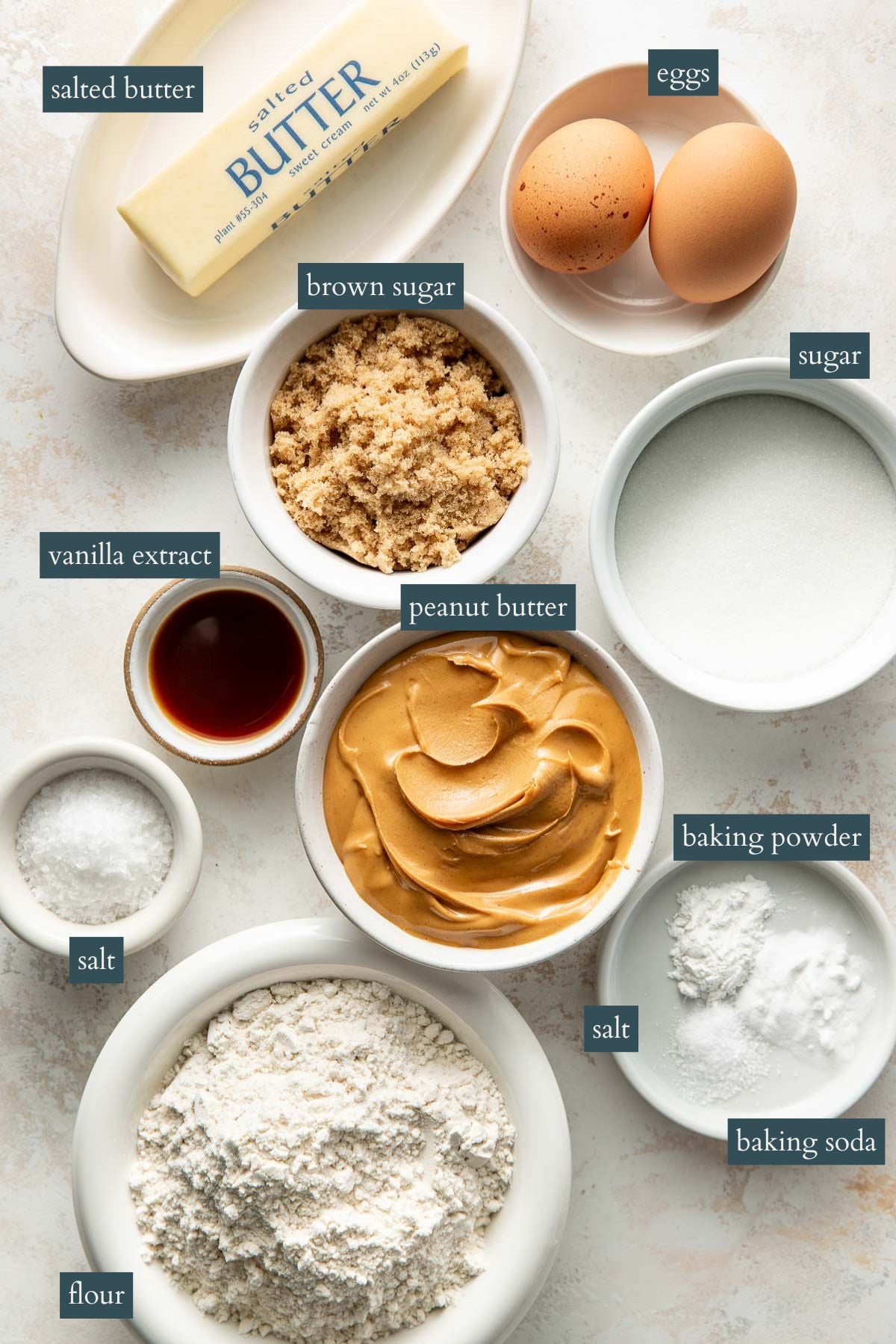 Various baking ingredients, including a stick of salted butter, two eggs, bowls of brown sugar, white sugar, vanilla extract, peanut butter, flour, baking powder, baking soda, and salt, arranged on a white surface.