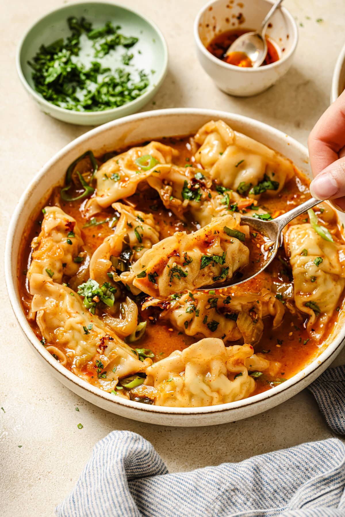 A hand holds a spoon over a bowl of dumplings in a spicy red broth, garnished with chopped herbs. Small bowls of fresh herbs and chili oil are in the background on a light surface.