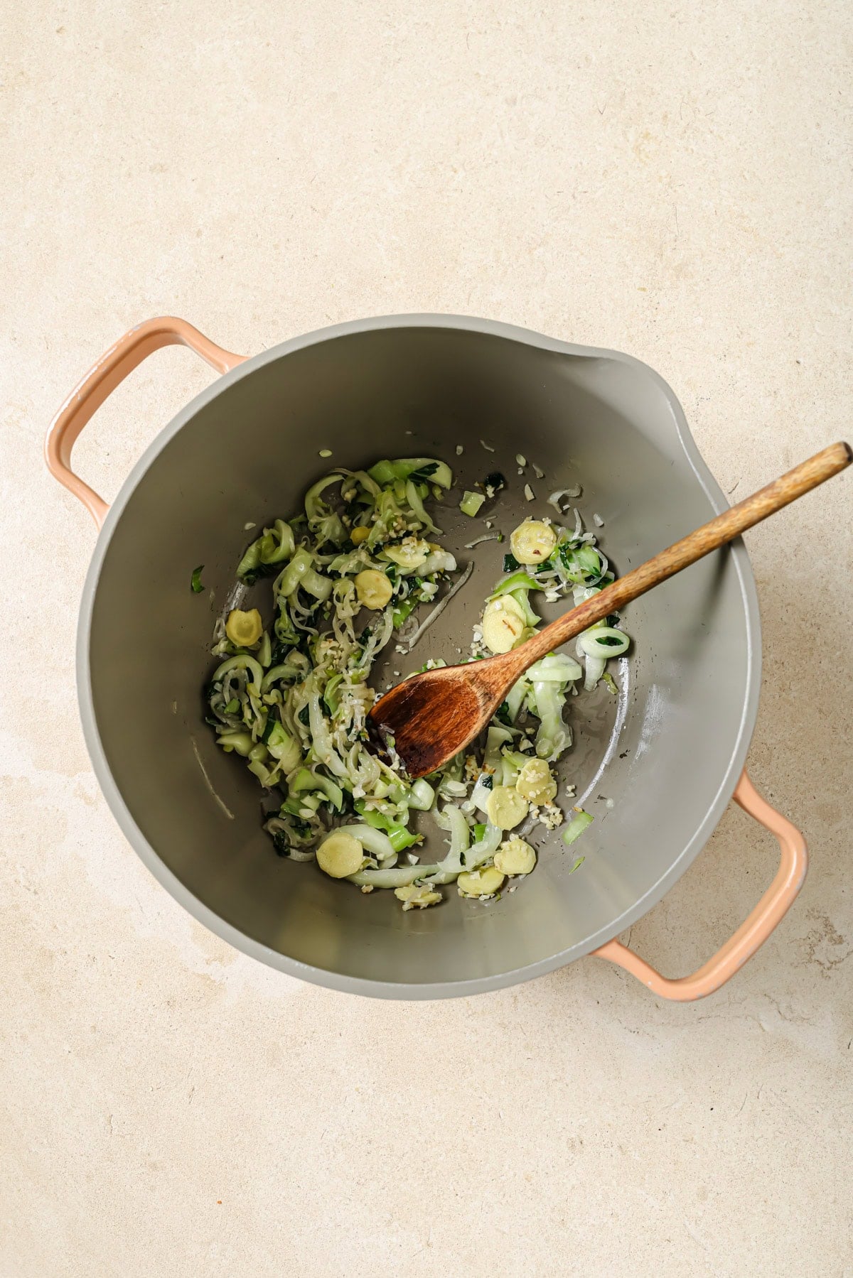 A top view of a pot with sautéed sliced zucchini, onions, and garlic, being stirred with a wooden spoon on a light-colored surface.