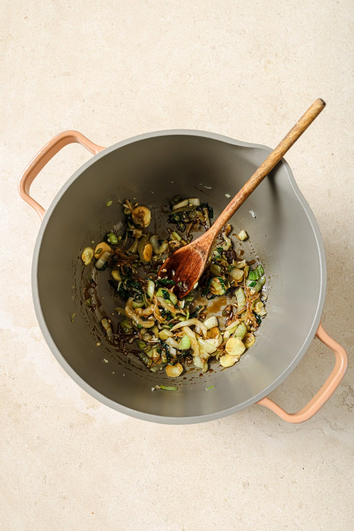 A beige pot with pink handles contains sautéed onions, mushrooms, and greens, being stirred with a wooden spoon, placed on a light-colored countertop.