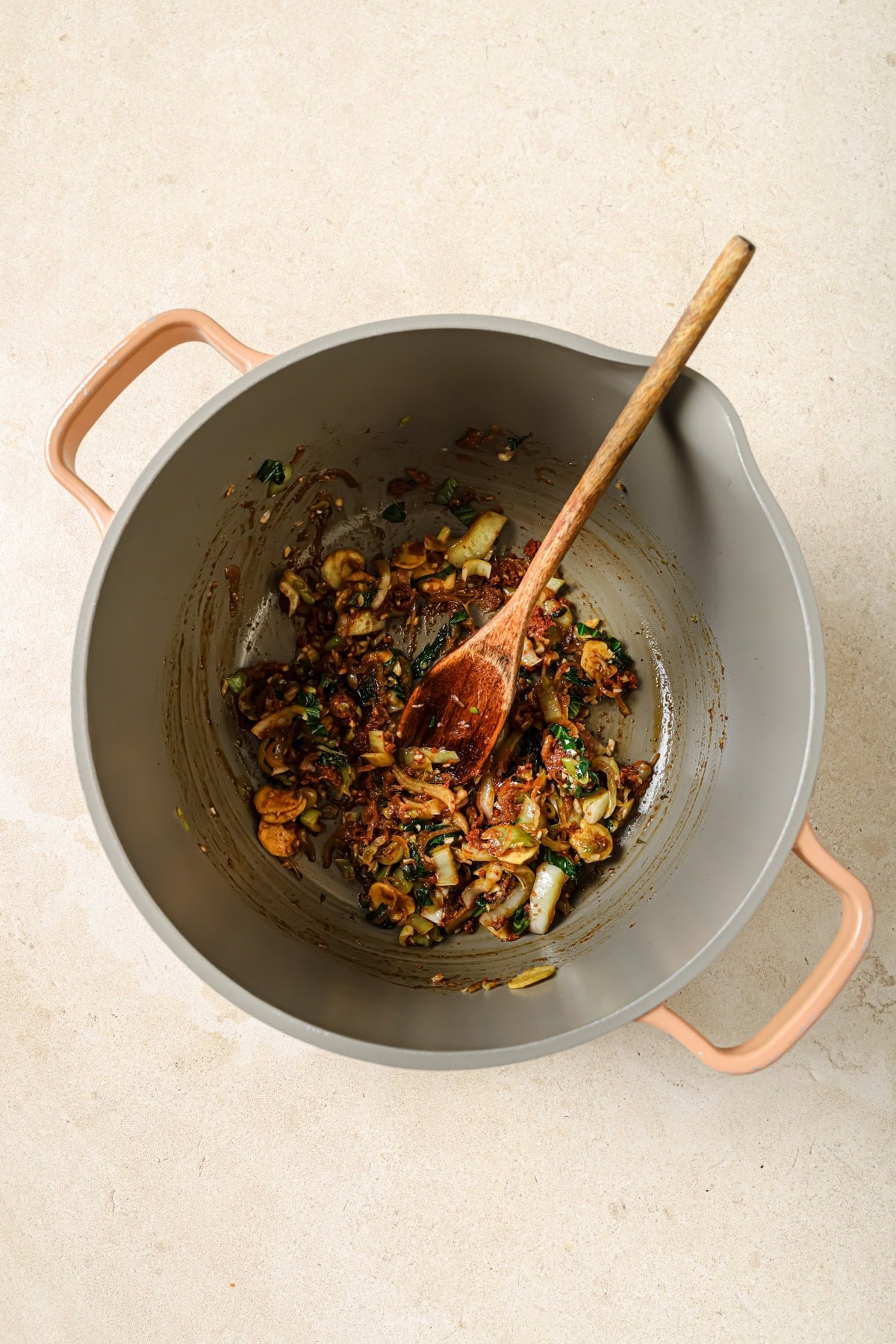 A gray pot with pink handles contains sautéed vegetables and herbs being stirred with a wooden spoon, sitting on a light-colored countertop.