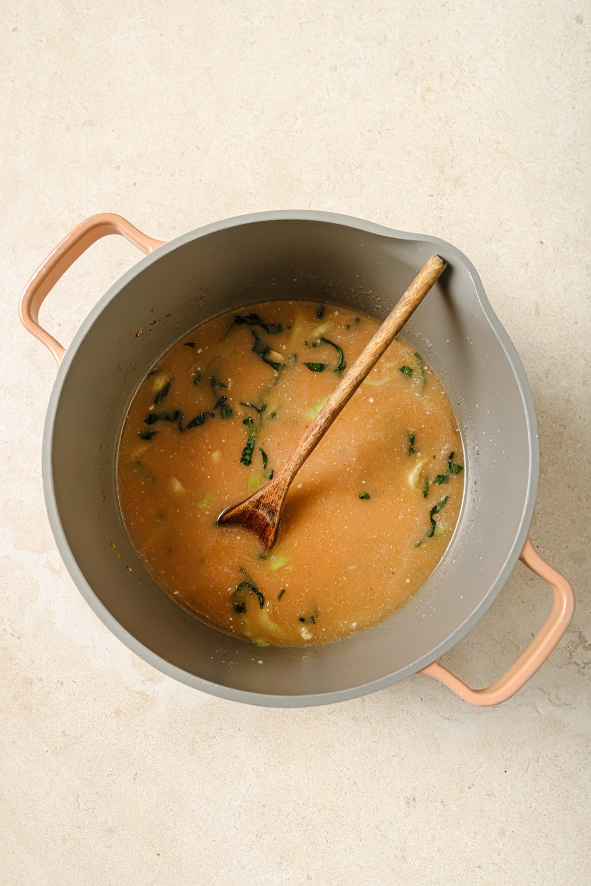 A large gray pot with peach-colored handles contains a light brown soup with leafy greens and a wooden spoon resting inside, set on a light beige surface.