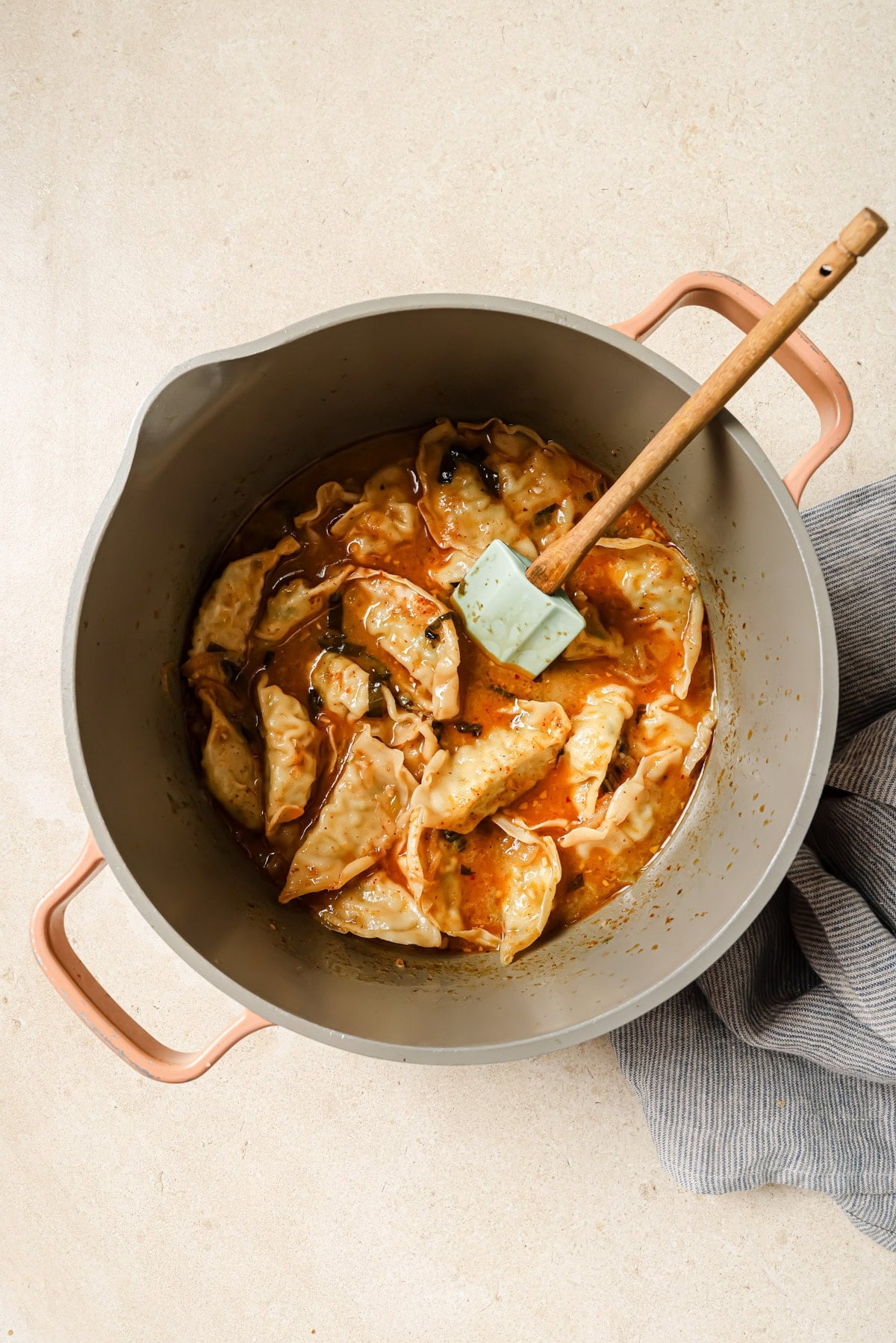 A pot filled with cooked dumplings in a reddish-orange sauce sits on a light-colored surface. A green spatula rests inside the pot, and a blue-striped cloth is partially visible nearby.