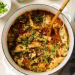 A white pot filled with dumplings in a spicy red broth, garnished with chopped herbs and green onions. A wooden spoon rests in the pot, with bowls of extra herbs and chili oil nearby on a light countertop.
