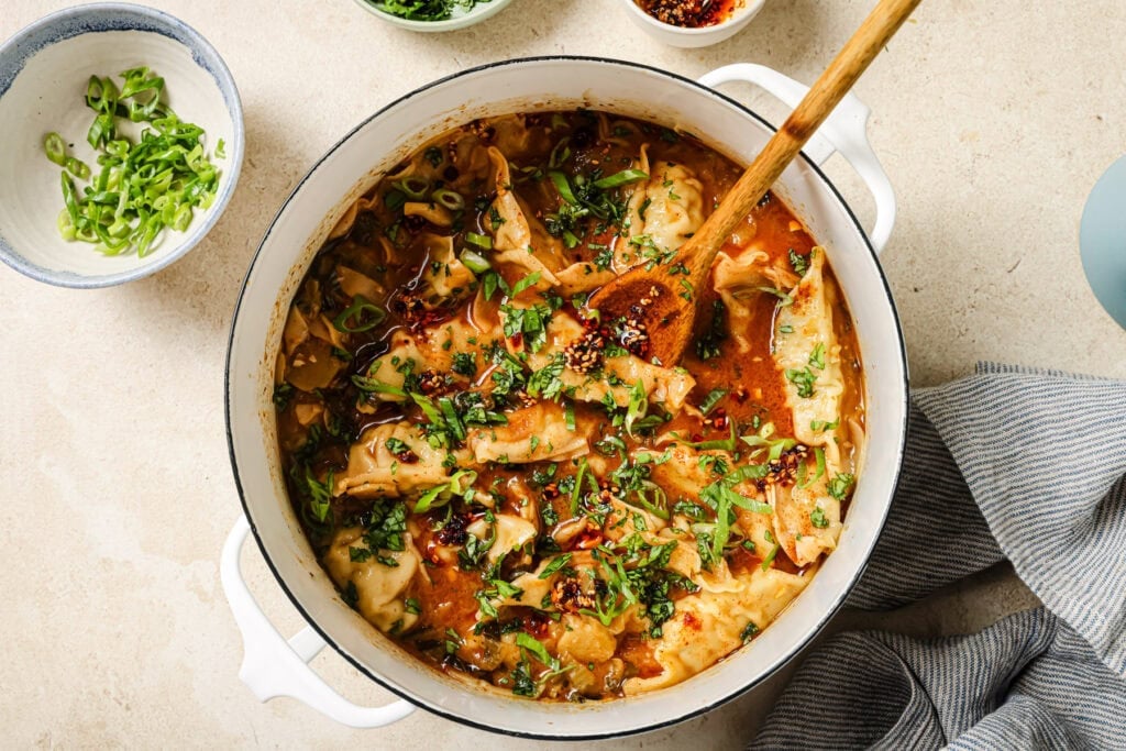 A white pot filled with dumplings in a spicy red broth, garnished with chopped green onions and herbs. A wooden spoon rests in the pot; bowls with extra toppings are nearby on a light surface.