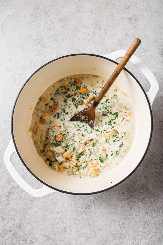 A white pot filled with a creamy soup containing mussels, chopped herbs, and onions, with a wooden spoon resting inside, on a light gray countertop.