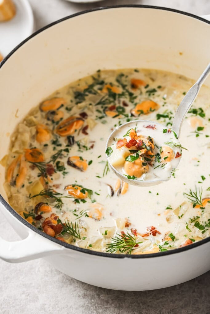 A pot of creamy seafood chowder garnished with fresh herbs, with mussels and bits of bacon visible. A ladle lifts a serving from the pot, showing the rich, chunky texture of the soup.