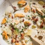 A bowl of creamy seafood chowder with mussels, diced potatoes, celery, bacon bits, fresh dill, and parsley, served with a slice of crusty bread and a spoon.