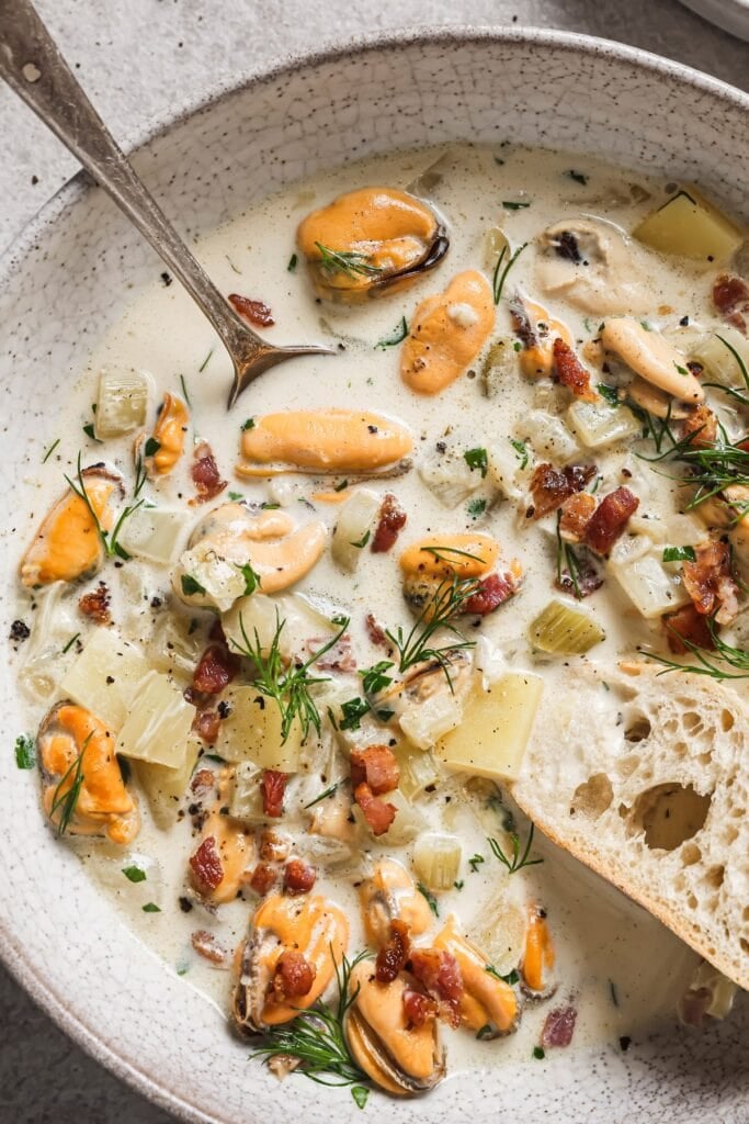 A bowl of creamy seafood chowder with mussels, diced potatoes, celery, bacon bits, fresh dill, and parsley, served with a slice of crusty bread and a spoon.