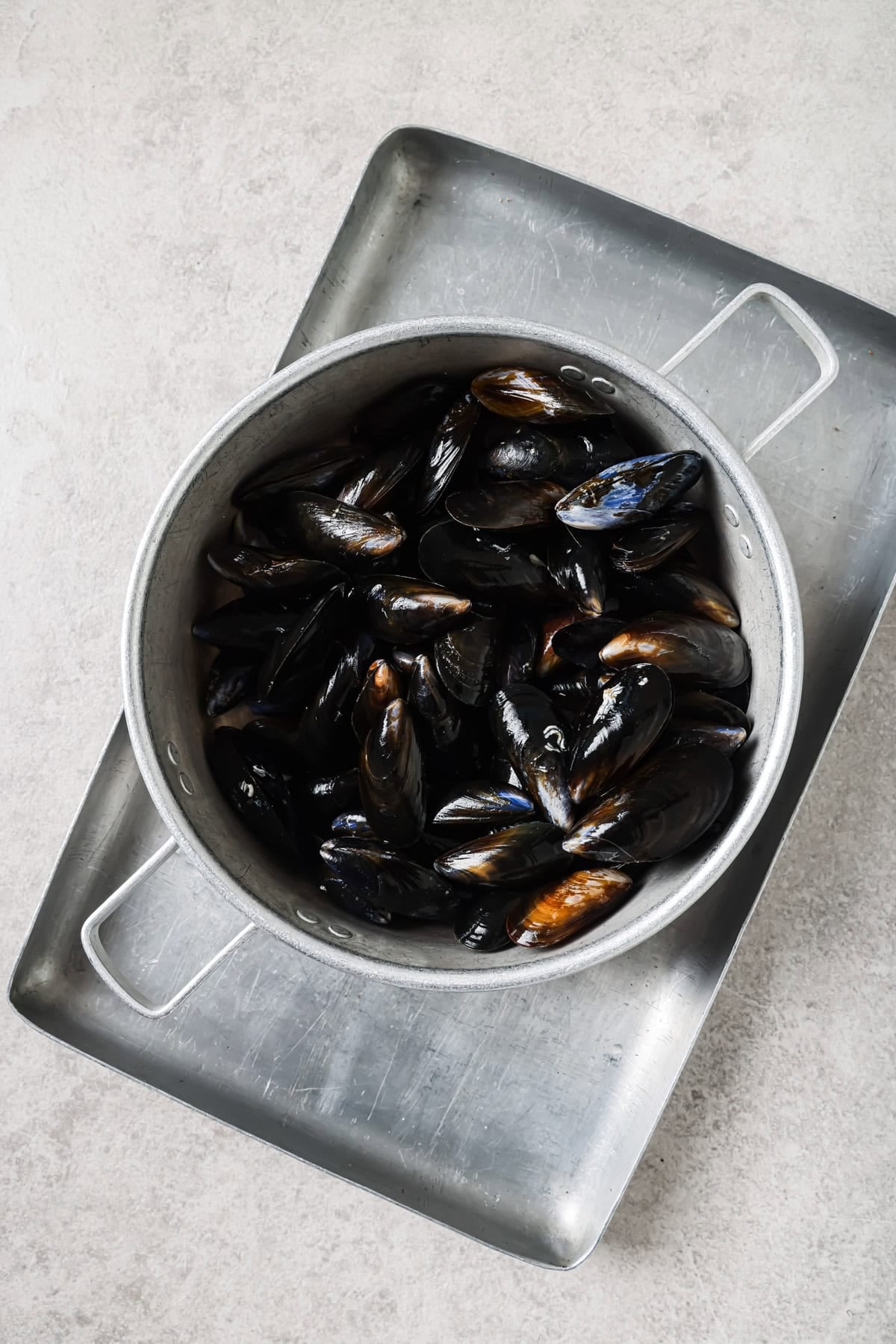 A large metal pot filled with cooked black mussels sits on a rectangular metal tray, placed on a light-colored textured surface.