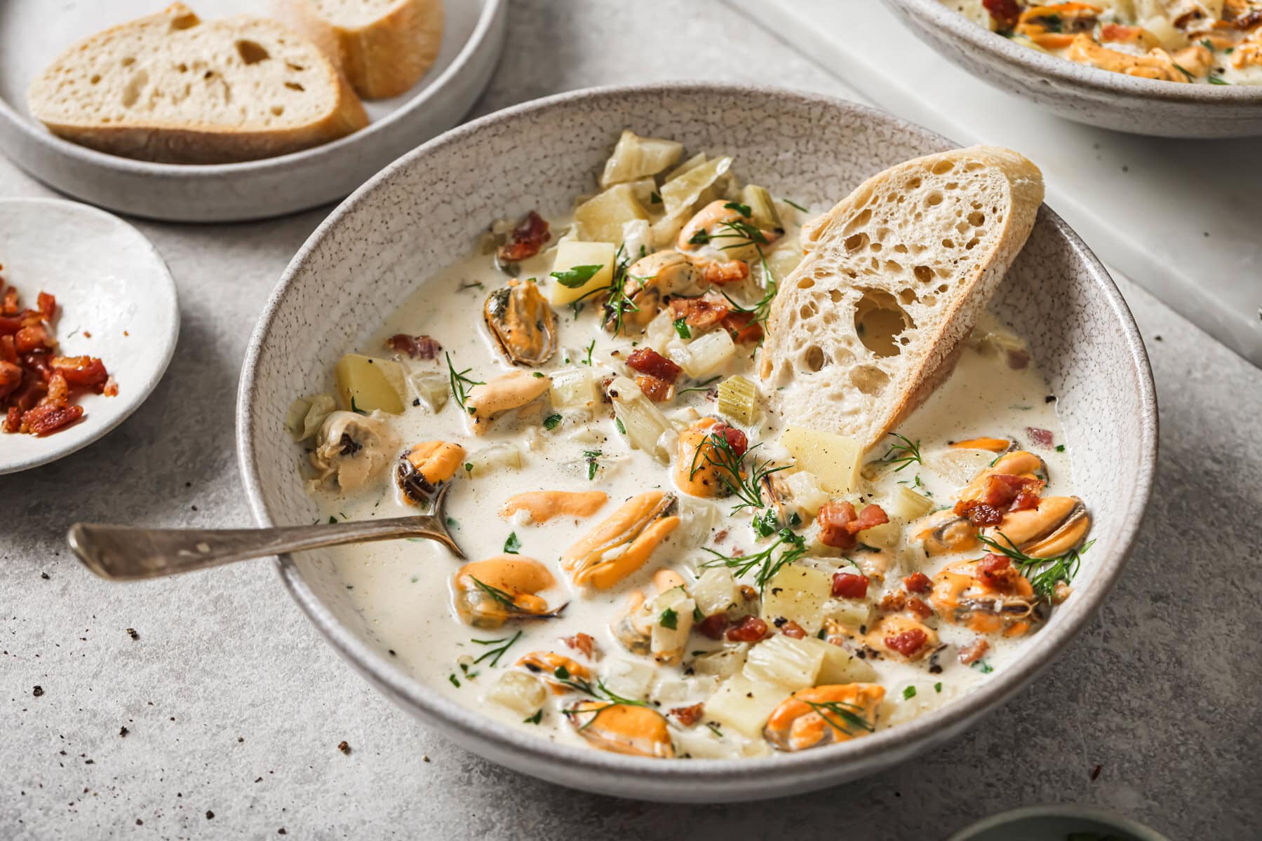 A bowl of creamy seafood chowder with mussels, diced potatoes, herbs, and bacon bits, served with a slice of crusty bread. A spoon rests in the bowl, and more bread is visible in the background.