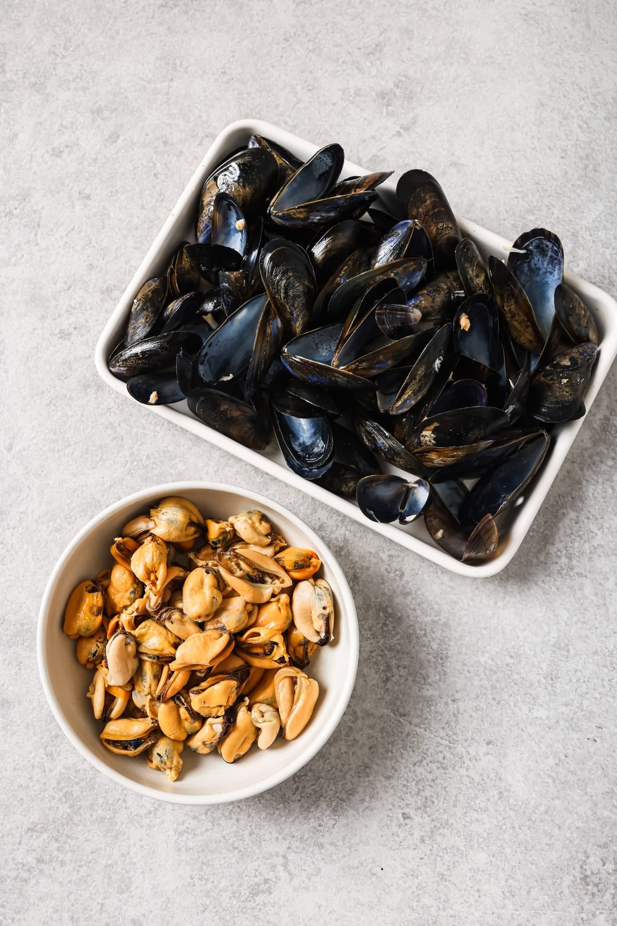 A rectangular dish with empty black mussel shells sits next to a round bowl filled with cooked, shelled mussel meat on a light gray surface.