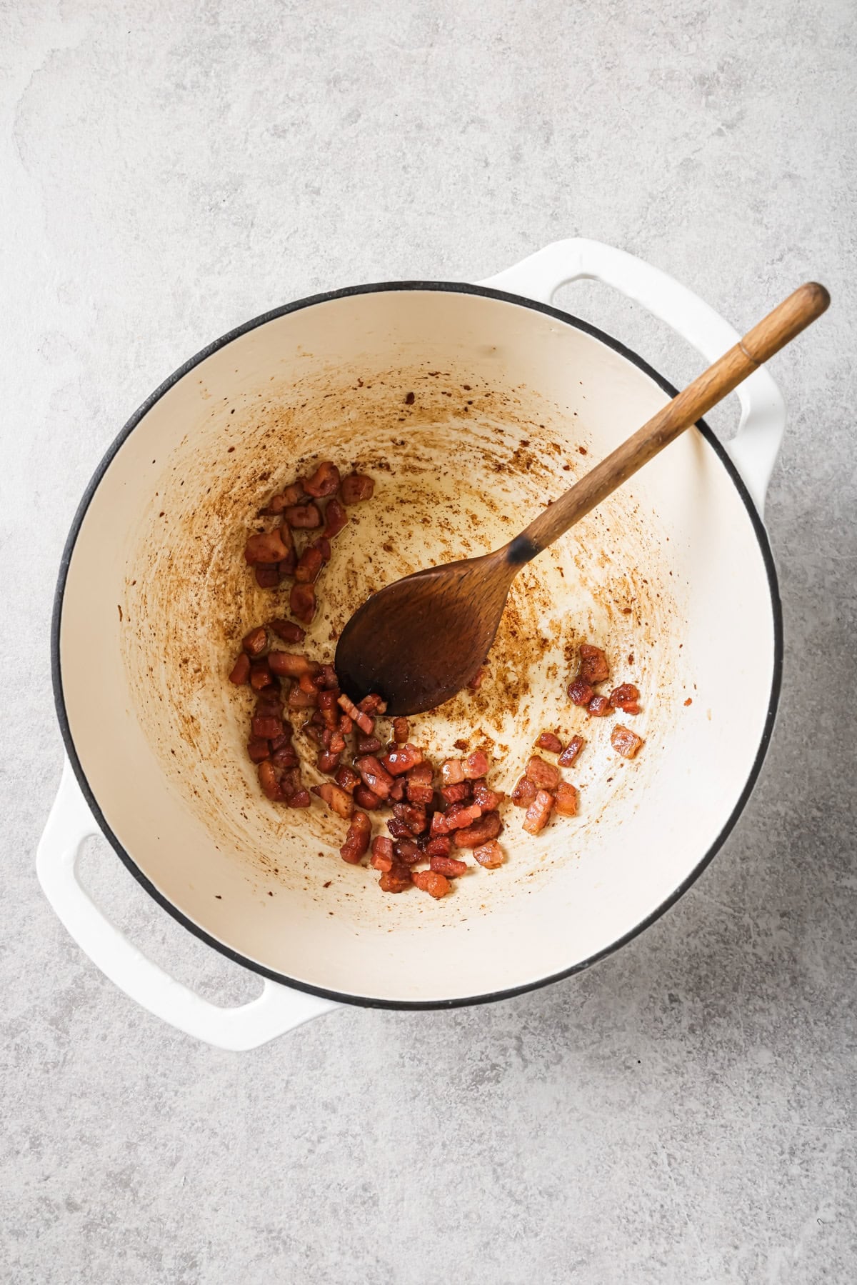Overhead view of a white Dutch oven with cooked bacon pieces and a wooden spoon resting inside, on a light gray countertop. The pot has browned bits and oil residue on the bottom.
