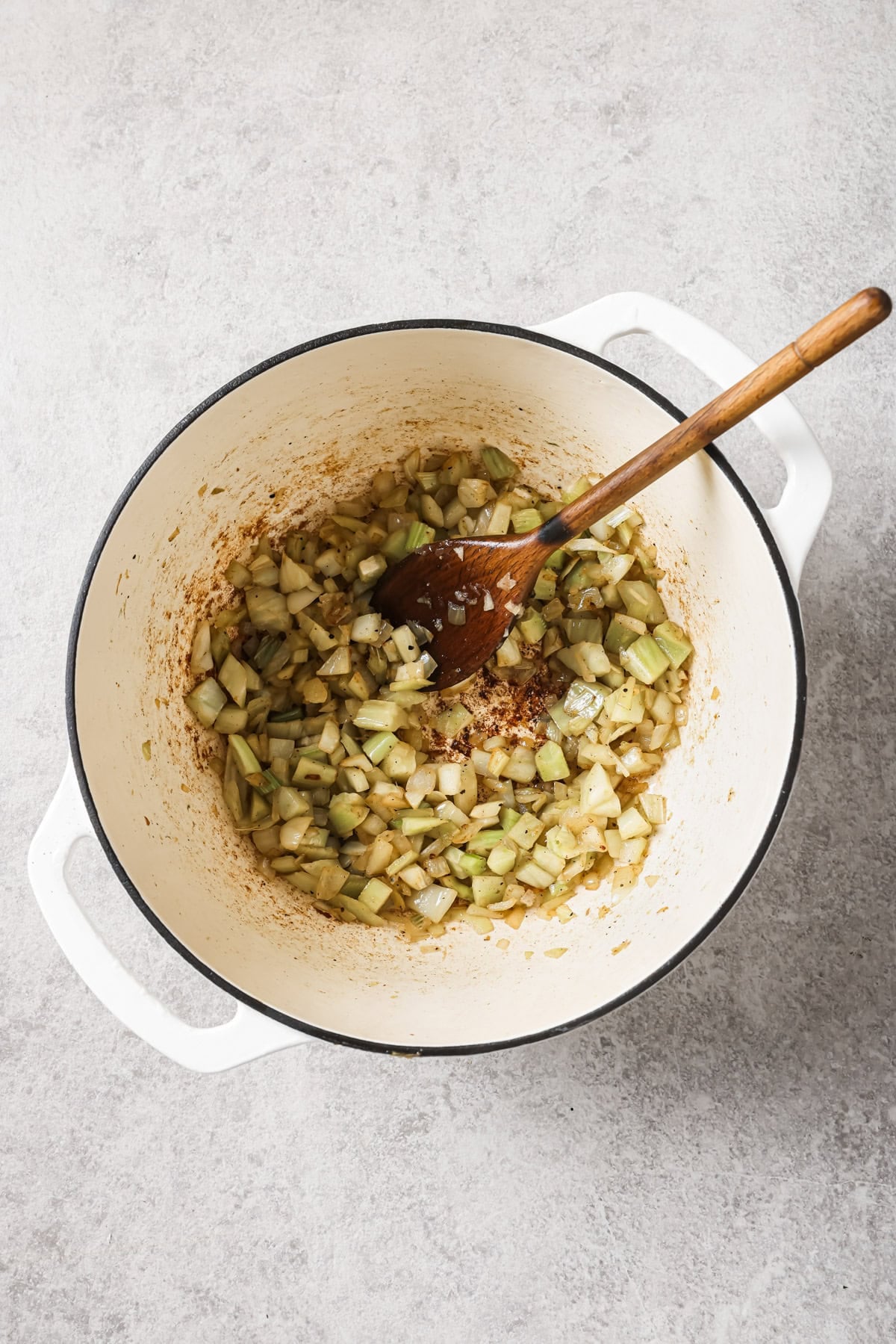 Diced onions and celery are being sautéed in a large white Dutch oven with a wooden spoon resting on the side. The mixture appears partially cooked and is lightly browned on the bottom.