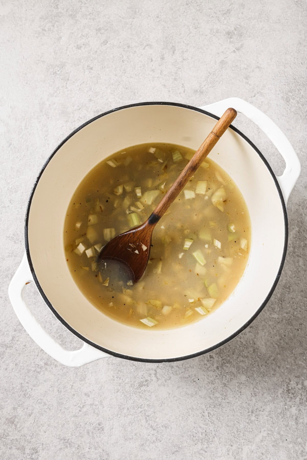 A white pot with handles contains a light broth with diced onions and vegetables, and a wooden spoon resting inside. The pot is placed on a light gray surface.