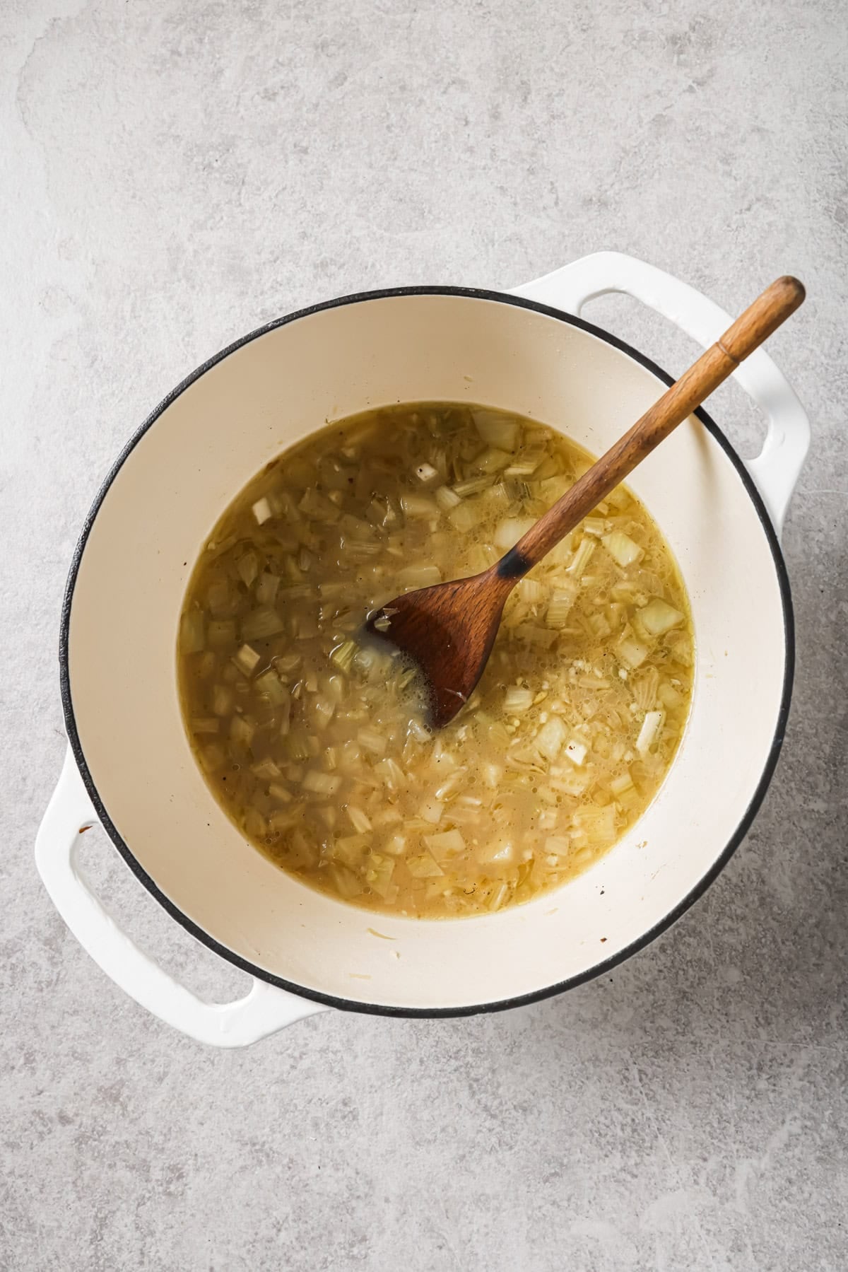 A white Dutch oven filled with chopped onions cooking in broth, stirred with a wooden spoon, sits on a light gray countertop.