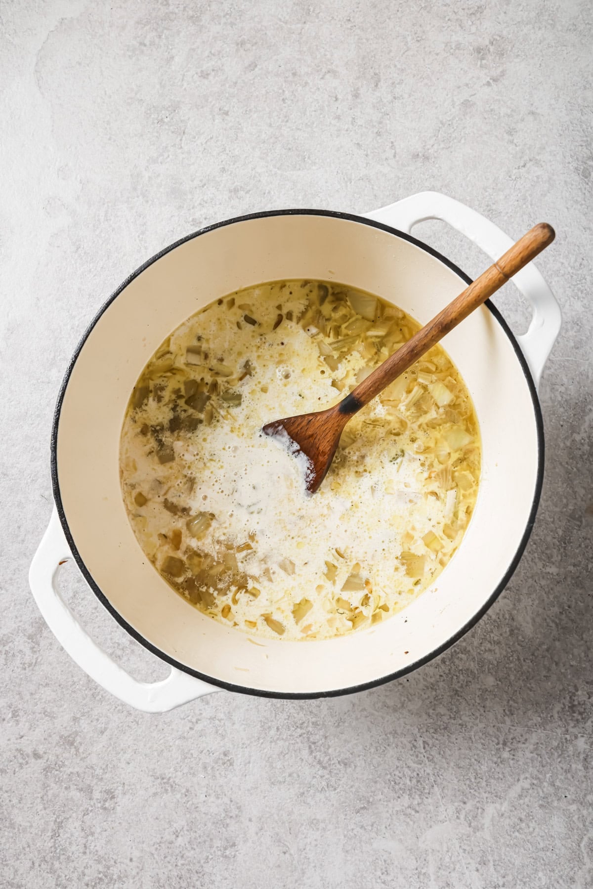 A white pot filled with soup, containing chopped vegetables and liquid, sits on a gray surface. A wooden spoon rests inside, and grated cheese is sprinkled on top.