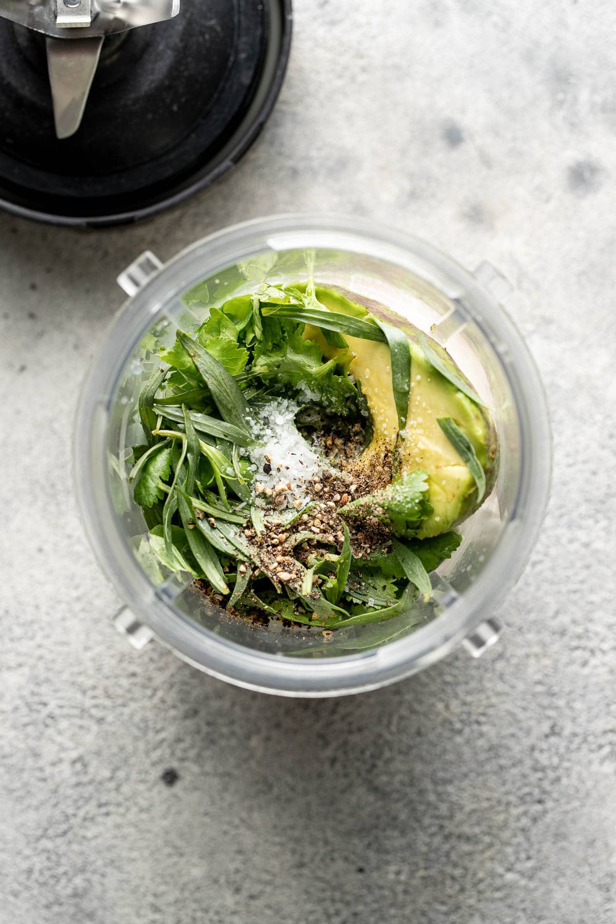 A blender cup filled with half an avocado, fresh green herbs, black pepper, and salt, ready to be blended, shown from above on a light gray surface.