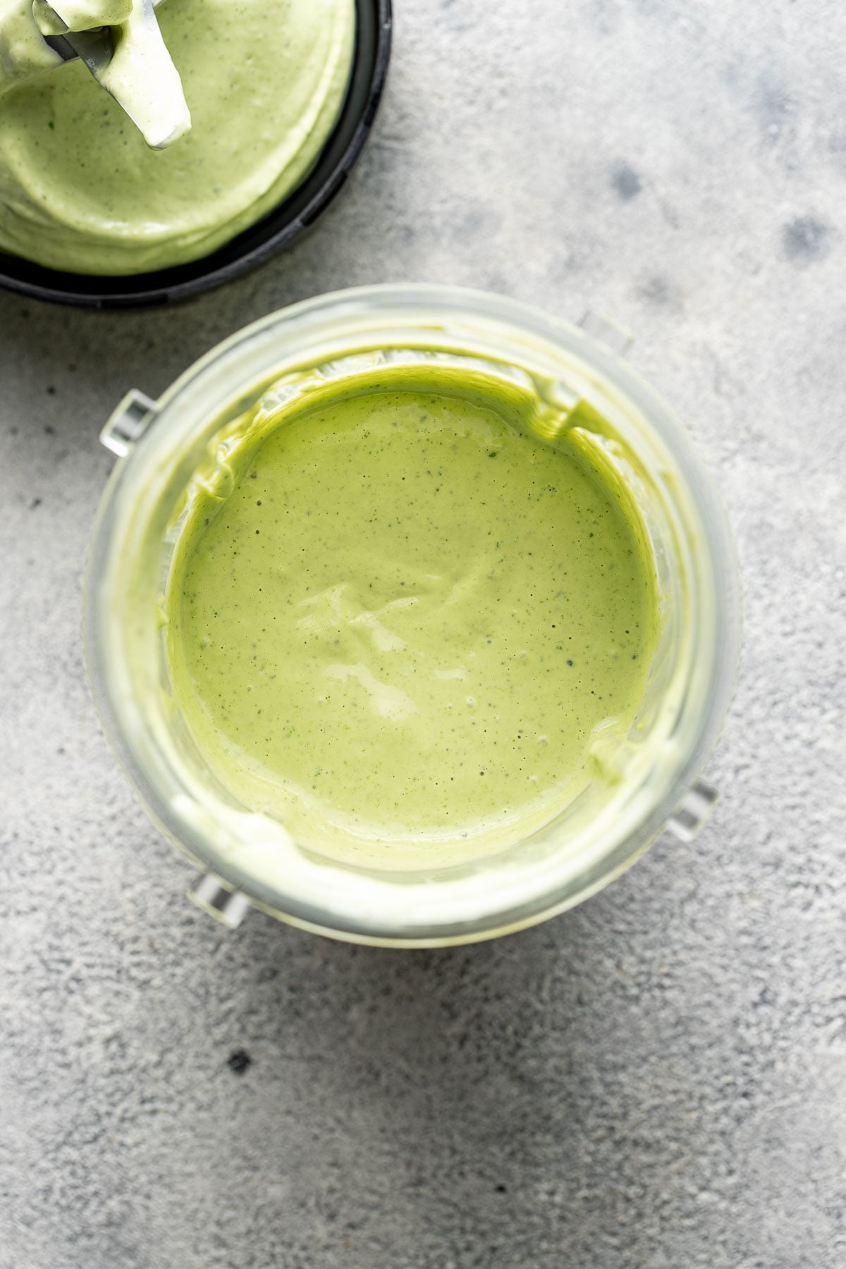 A blender filled with creamy green sauce sits on a light gray surface, with the detached blade assembly visible nearby.