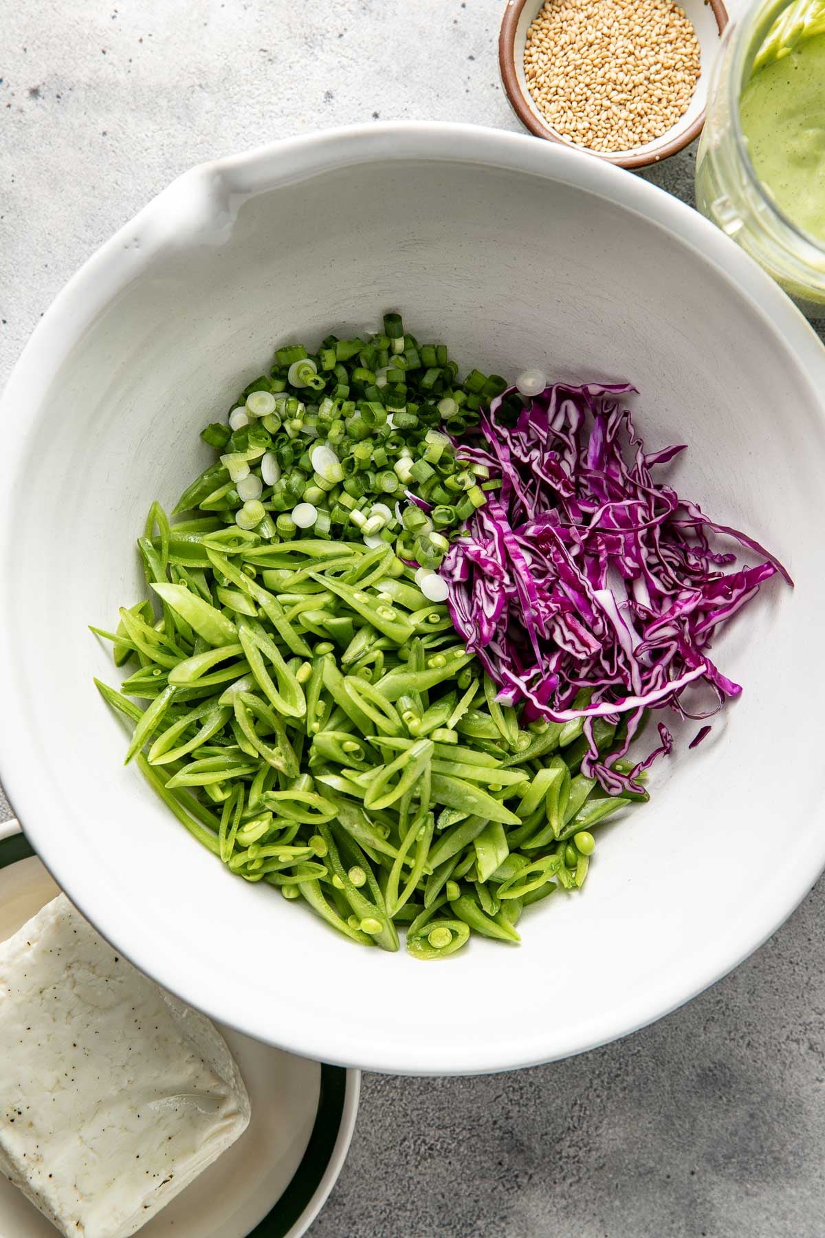 A white bowl containing sliced snap peas, shredded purple cabbage, and chopped green onions, with small bowls of sesame seeds and a block of tofu nearby.