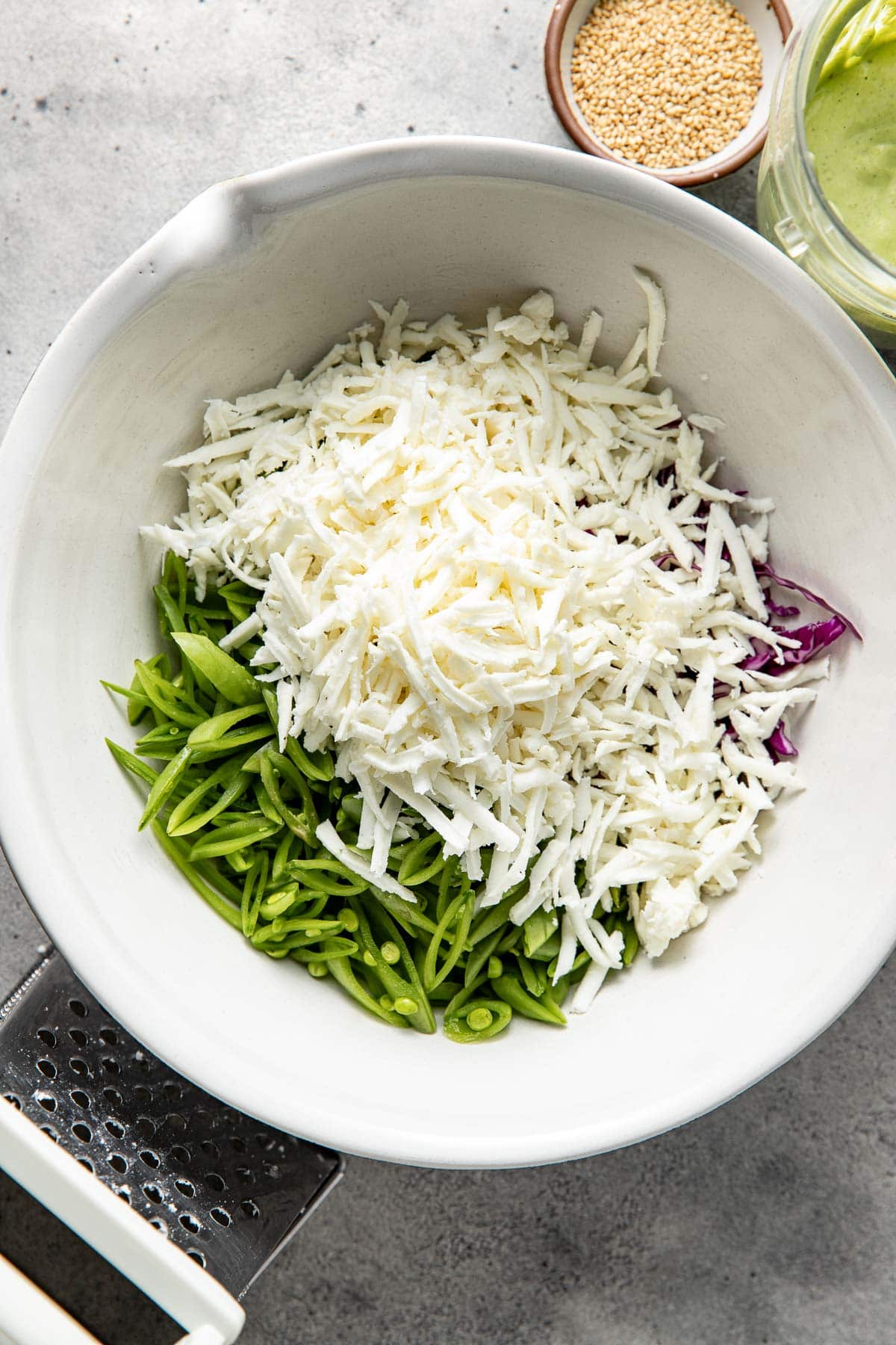 A white bowl contains shredded white cheese, sliced green snap peas, and purple cabbage. Nearby are a small bowl of sesame seeds and a jar of green dressing on a gray surface.