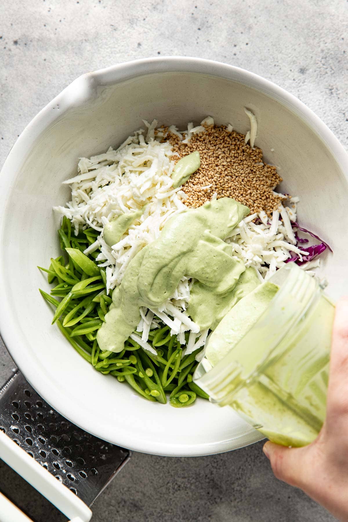A white bowl filled with sliced snap peas, shredded cabbage, grated coconut, sesame seeds, and a creamy green dressing being poured on top.