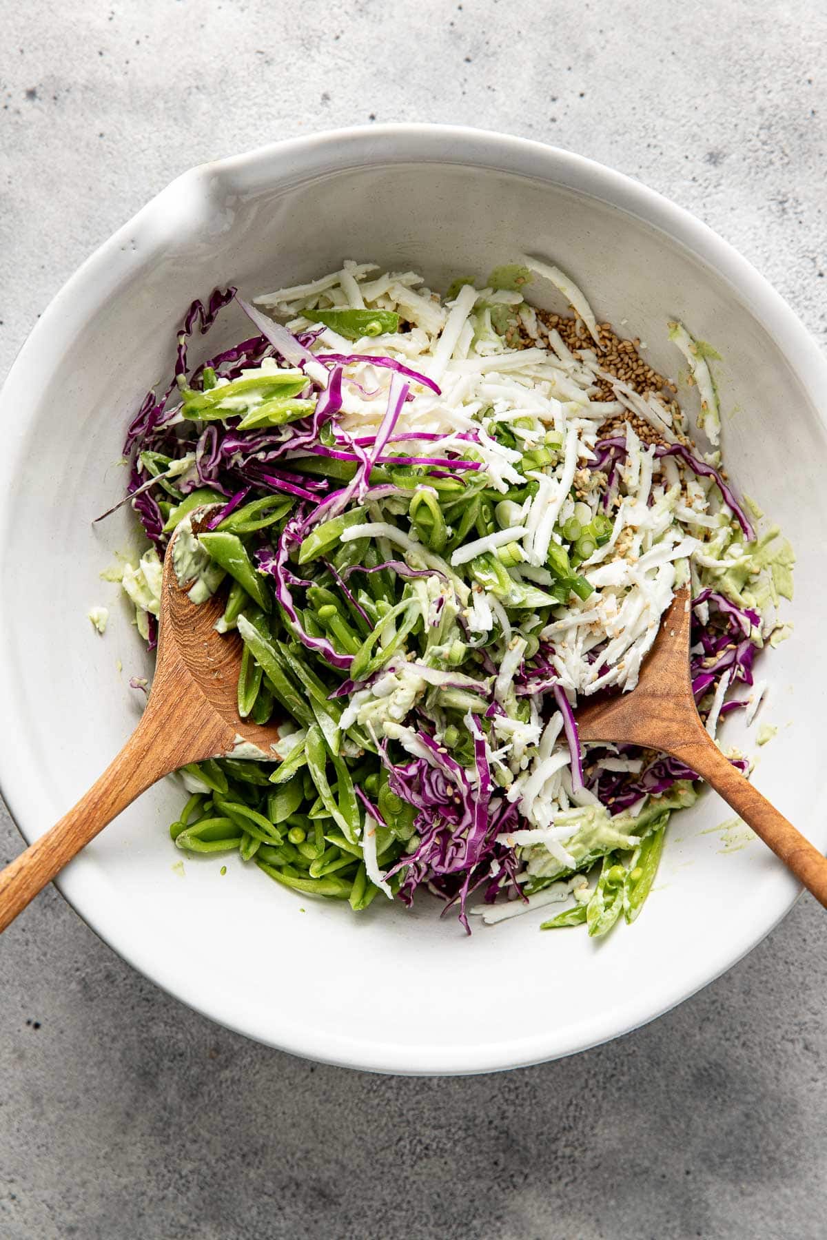 A white bowl contains shredded green and purple cabbage, green snow peas, and sesame seeds. Two wooden salad servers rest inside the bowl on a light gray surface.