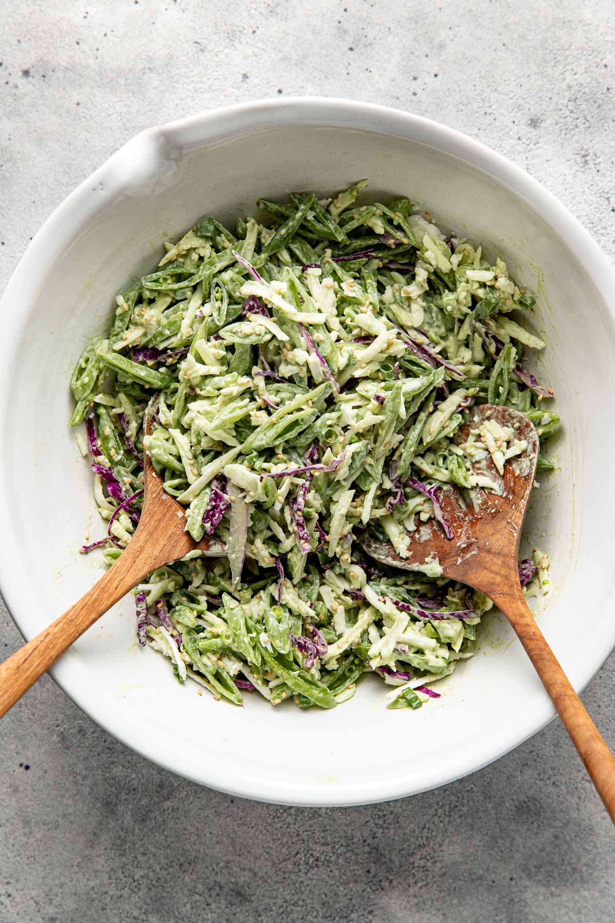A large white bowl filled with creamy coleslaw made of shredded cabbage and green vegetables, mixed with dressing. Two wooden serving spoons are placed inside the bowl.