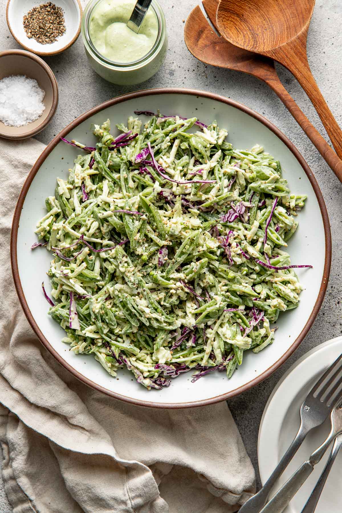 A large bowl of shredded green and purple cabbage salad with a creamy green dressing, surrounded by wooden utensils, plates, a jar of dressing, and small bowls of salt and pepper on a light surface.