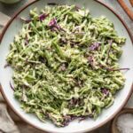 A plate of creamy broccoli slaw with shredded broccoli stems, red cabbage, and a green dressing, garnished with seeds. Wooden utensils, a jar of dressing, and small bowls of seasoning are nearby.