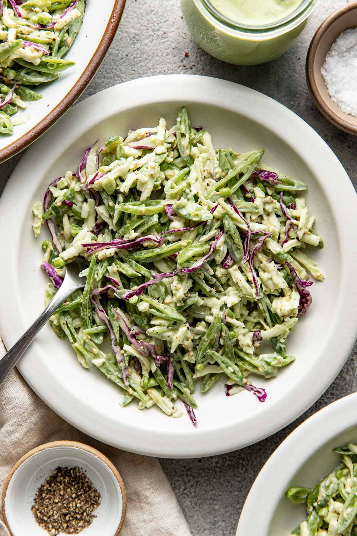 A white plate filled with creamy coleslaw made of shredded green beans, cabbage, and a light dressing, with a fork resting on the plate. Small bowls of salt, pepper, and dressing are nearby.