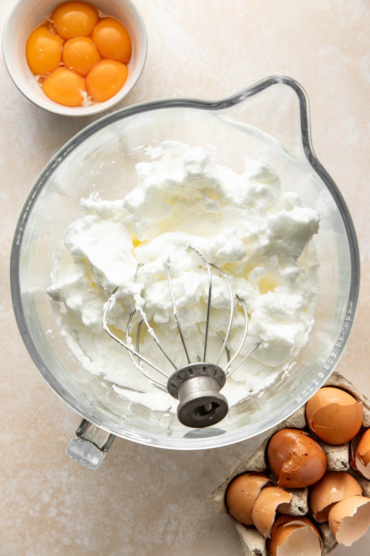 A mixing bowl with whipped egg whites and a whisk attachment, a small bowl with egg yolks, and an egg carton holding cracked brown eggshells on a light countertop.
