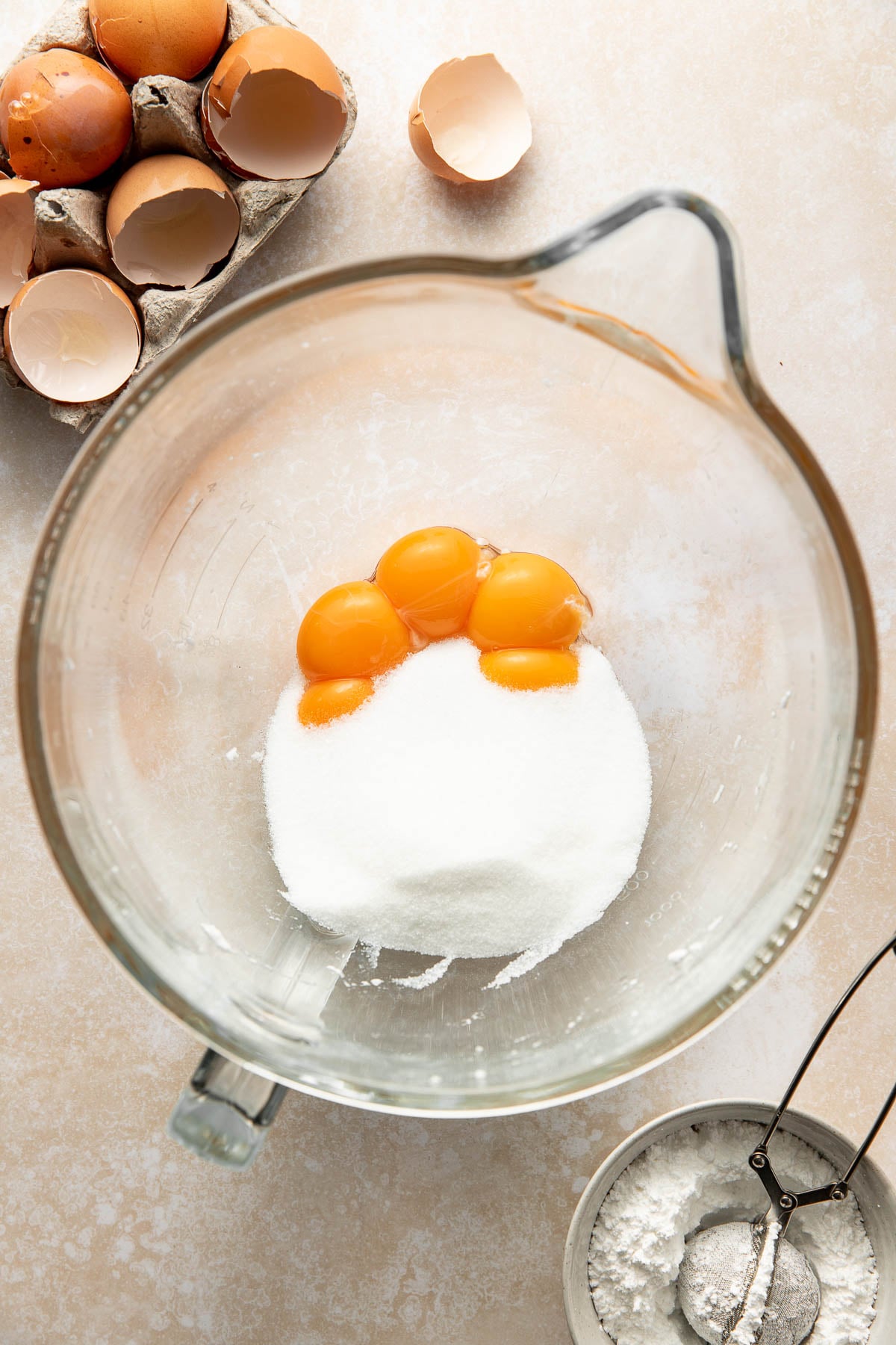A glass mixing bowl contains four egg yolks and a mound of granulated sugar. Nearby are cracked egg shells in a carton and a small bowl of flour with a sifter. All items rest on a light-colored surface.