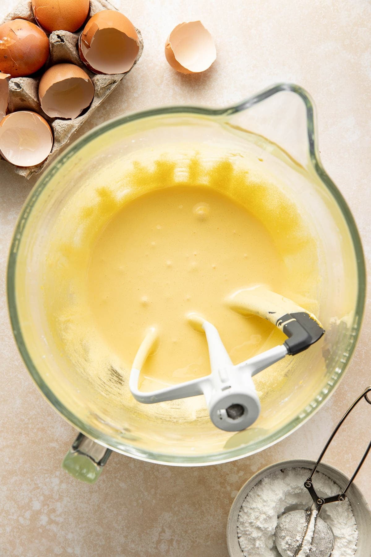 A glass mixing bowl with yellow cake batter and a stand mixer attachment, surrounded by cracked eggshells in a carton and a small bowl of flour with a sifter on a light countertop.