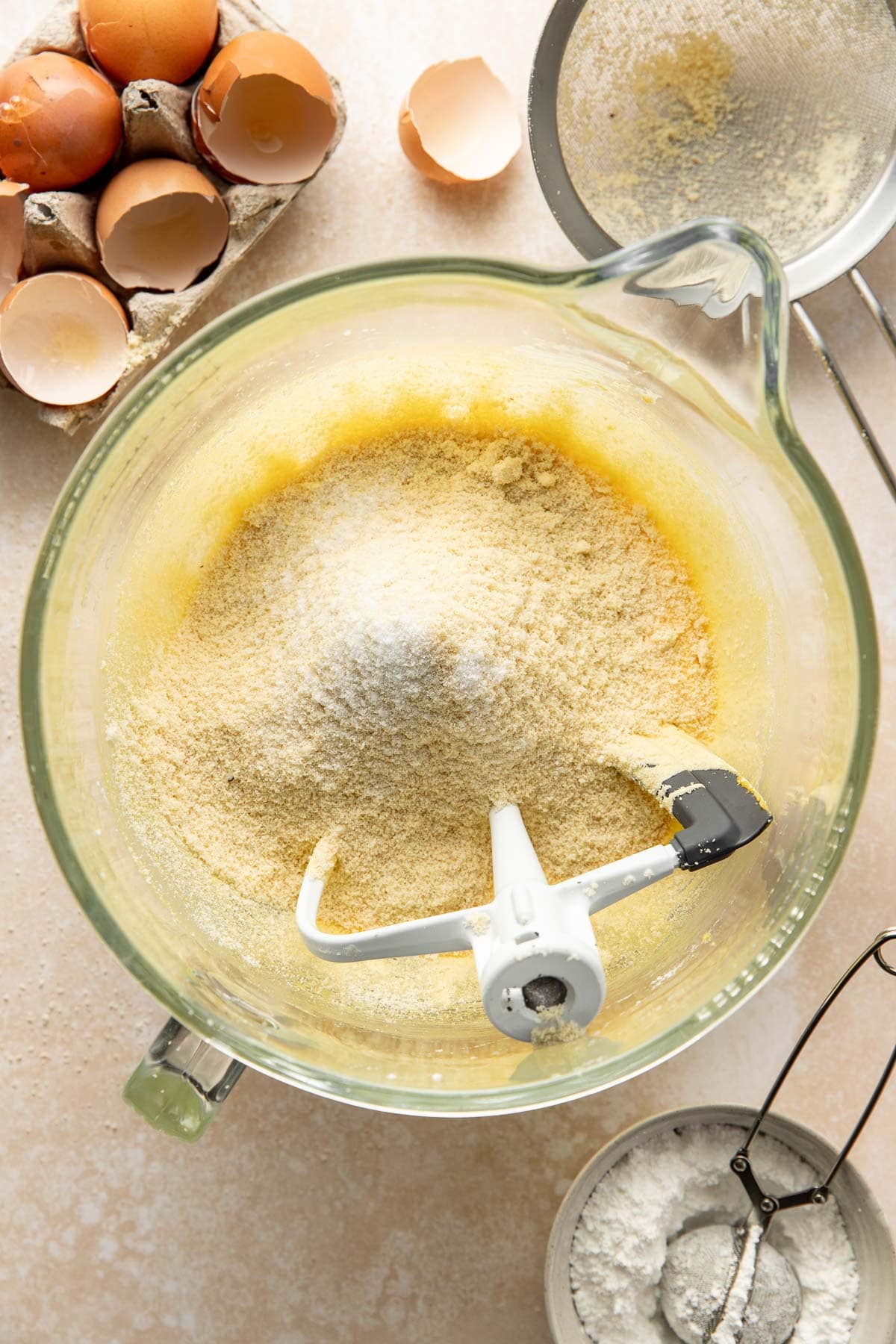 A glass mixing bowl containing cake batter and dry ingredients sits on a countertop, with a stand mixer paddle attached. Surrounding the bowl are cracked eggshells, a sifter with flour, and a bowl of flour.